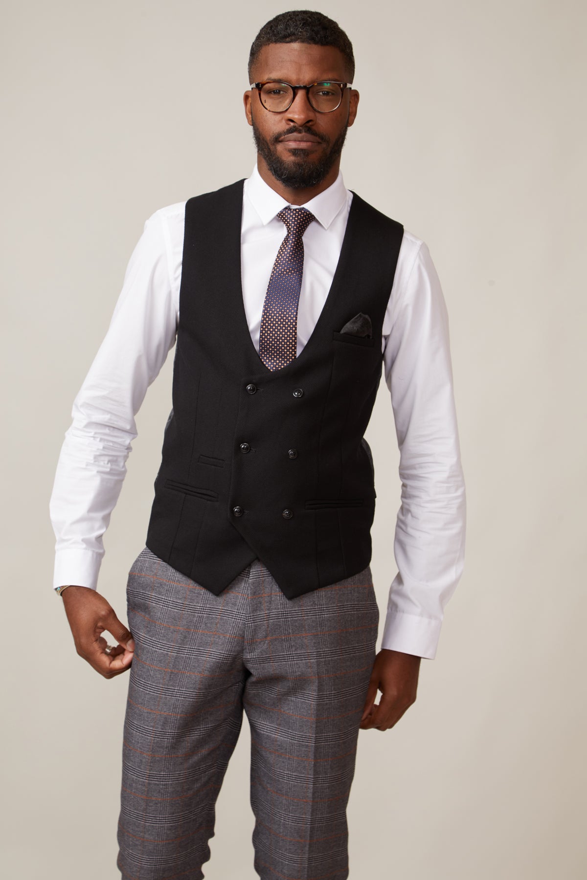 A man wearing glasses, a white dress shirt, marcdarcys KELVIN black double breasted waistcoat with paisley print lining, a patterned tie, and gray plaid trousers stands against a plain light background.