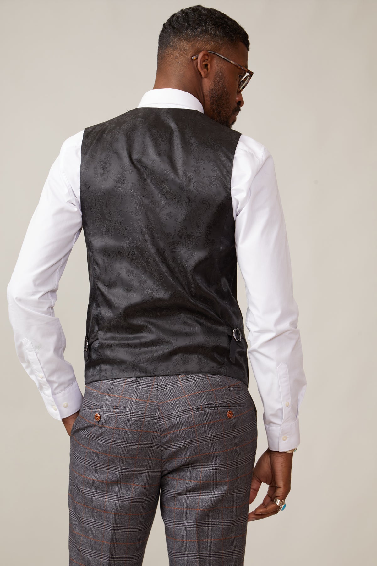 A man with short hair, glasses, and a ring faces away from the camera, wearing a marcdarcy KELVIN black double-breasted waistcoat, a white shirt, and gray plaid trousers with brown buttons.