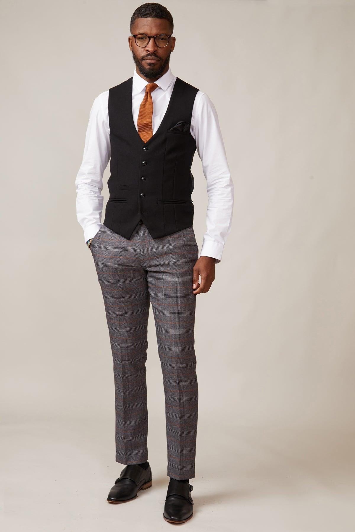 A man stands confidently in glasses, a white dress shirt, slim fit gray plaid pants, an orange-brown tie, black shoes, and the marcdarcy KELVIN Black Single Breasted Waistcoat with paisley print lining against a light background.