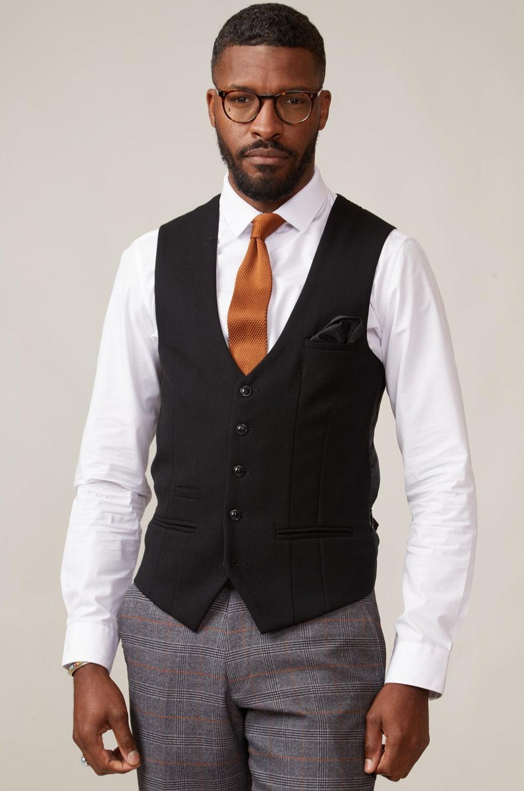 A man wearing glasses, a white shirt, an orange tie, plaid trousers, and the marcdarcy KELVIN black single breasted waistcoat stands facing forward against a plain background.