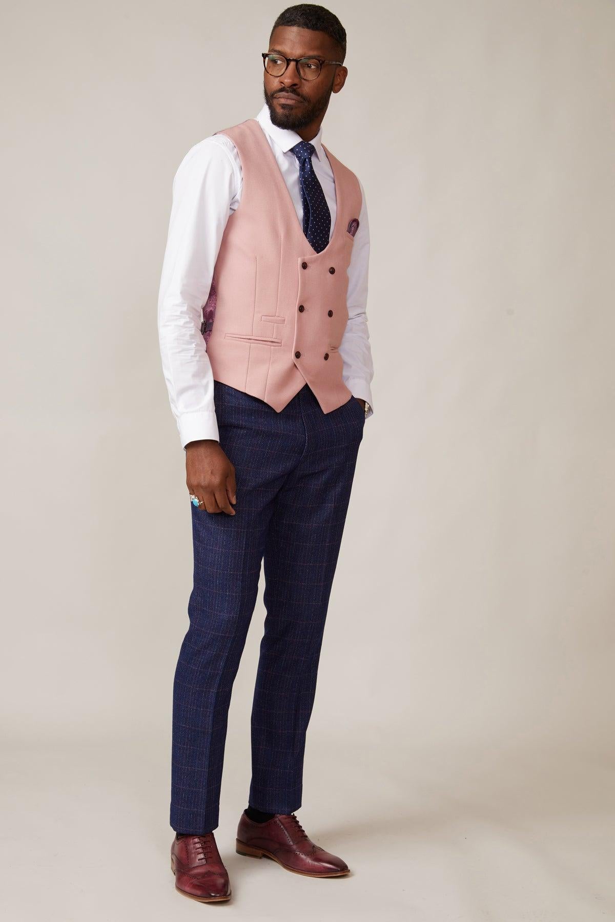 A man wearing the marcdarcy KELVIN Pink Double Breasted Waistcoat over a white dress shirt, navy checkered trousers, navy tie, and burgundy dress shoes stands against a plain light background, gazing to the side.