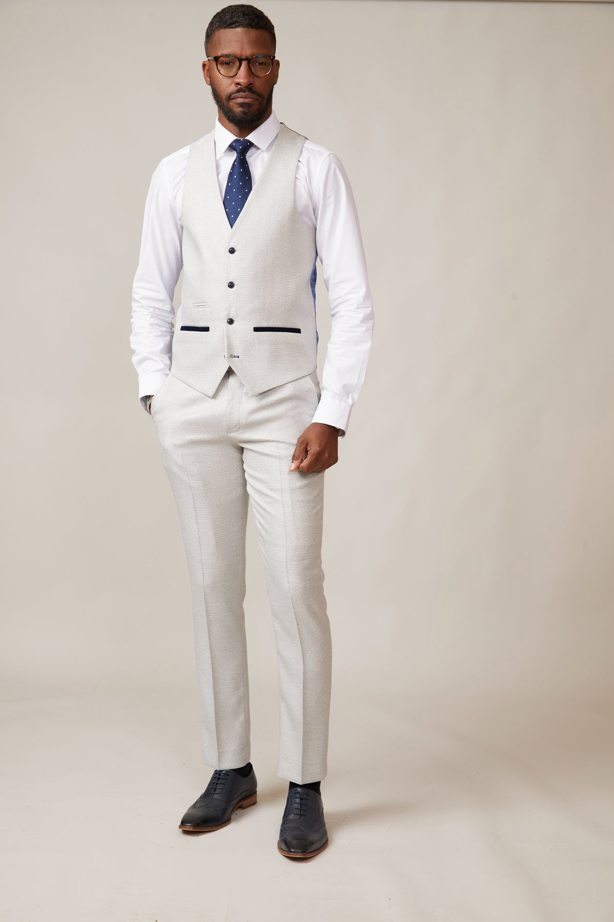 A man stands against a plain background wearing the marcdarcy RONALD Stone Single Breasted Waistcoat with matching trousers, a white dress shirt, navy polka-dot tie, black dress shoes, and glasses.