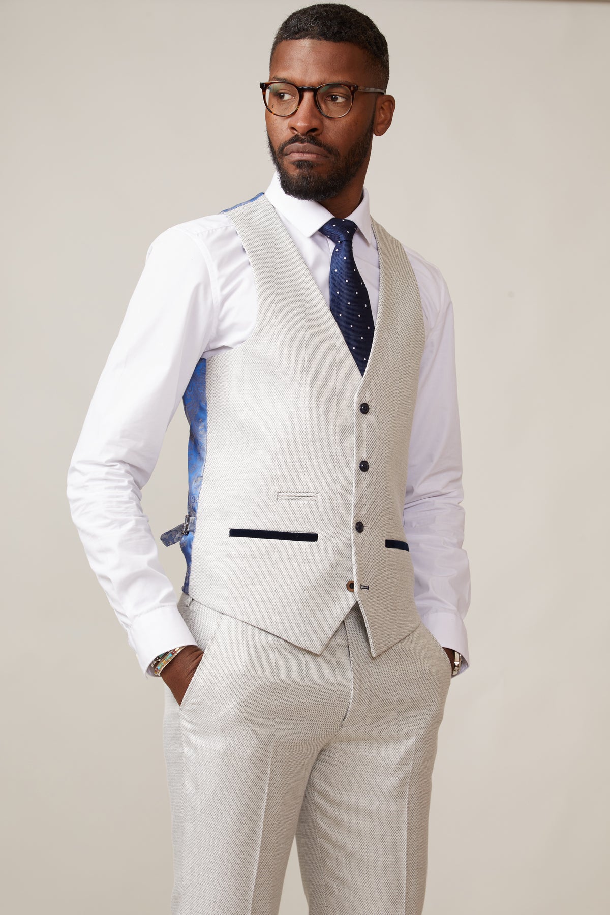 A bearded man in glasses wears the marcdarcy RONALD Stone Single Breasted Waistcoat as part of a slim fit light grey three-piece suit, styled with a white penny collar shirt and dark blue polka-dot tie, standing hands in pockets against a plain background.