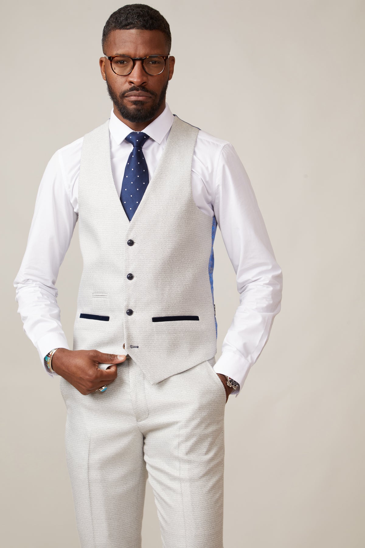 A man wears the marcdarcy RONALD Stone Single Breasted Waistcoat over a white penny collar shirt with a navy polka dot tie. He has glasses and his hands are in his pockets.