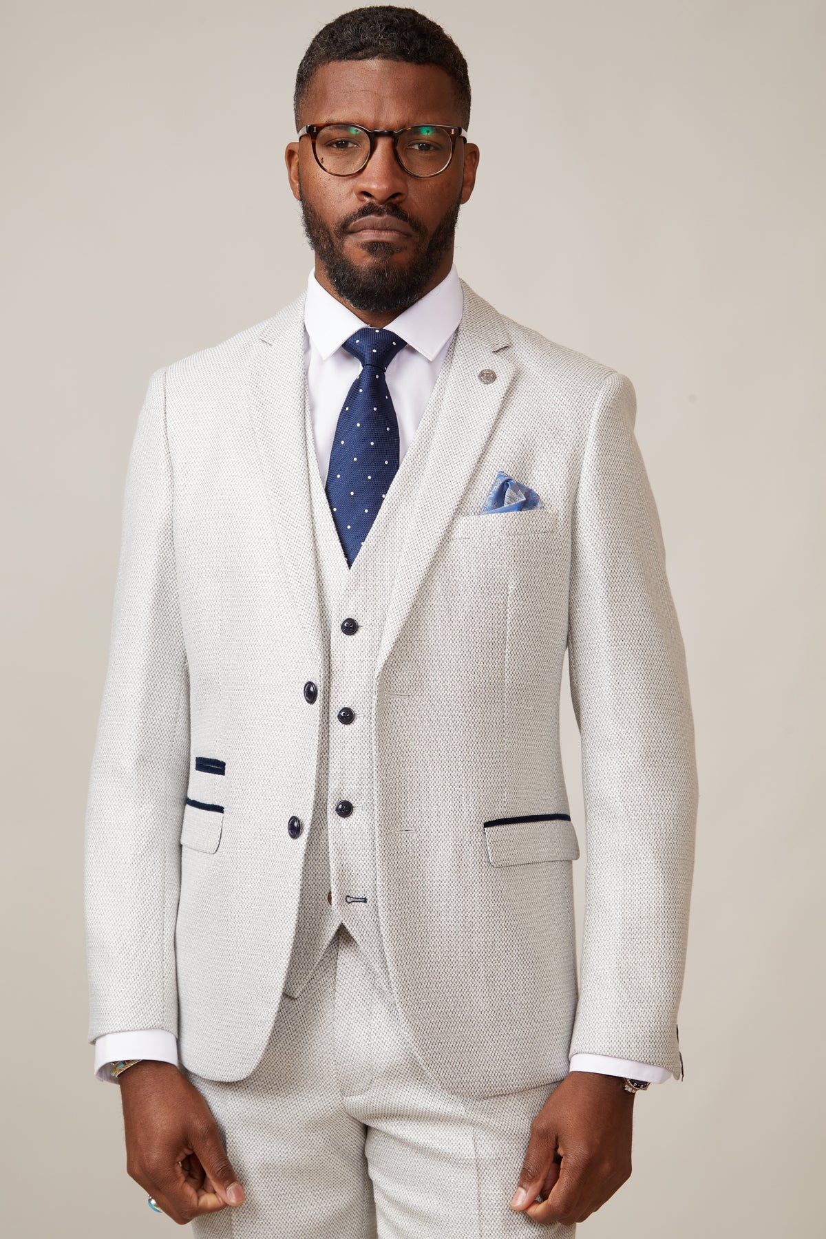 A bearded man with glasses, wearing a marcdarcy RONALD - Stone Tailored Blazer with paisley lining, white shirt, navy polka dot tie, and blue pocket square stands against a beige background with a serious expression.