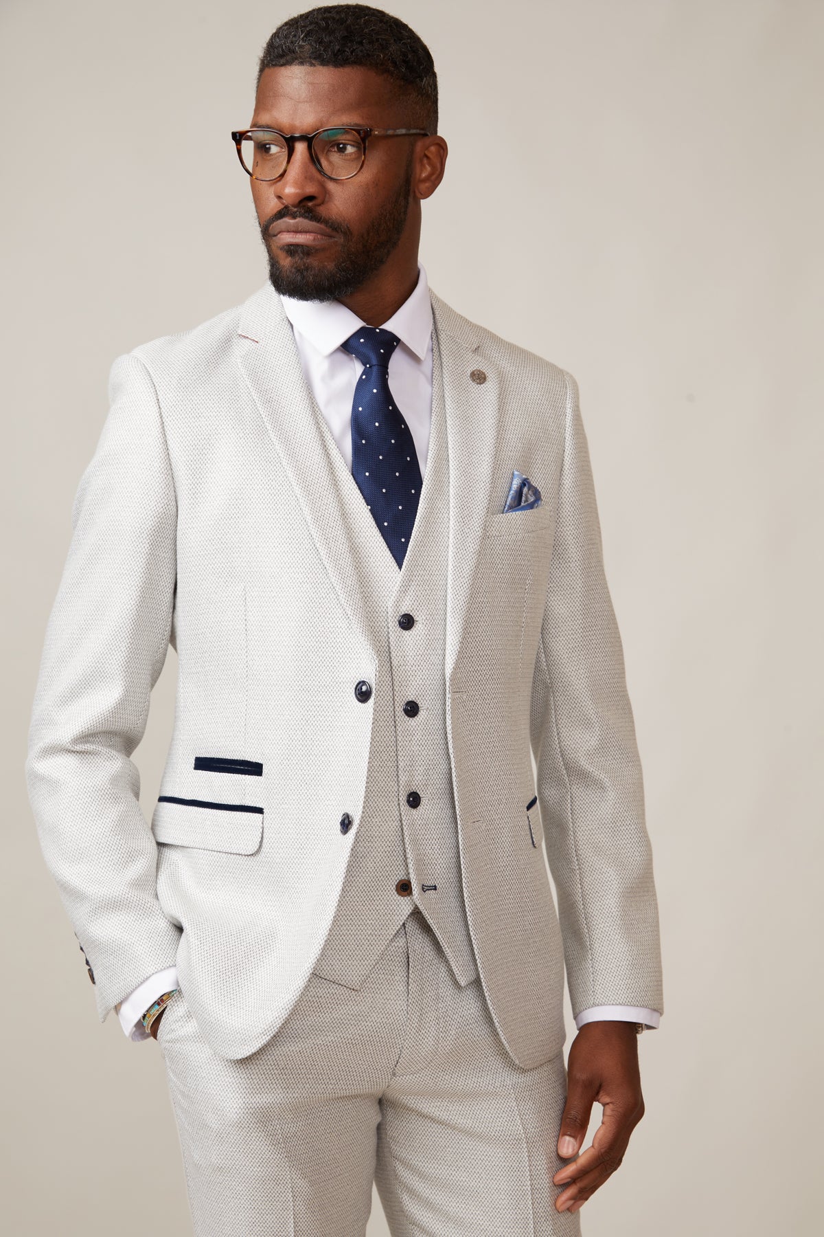 A Black man in glasses wears the marcdarcy RONALD - Stone Tailored Blazer as part of a three-piece suit, styled with a dark blue polka dot tie and pocket square, standing confidently with his hand in his pocket.