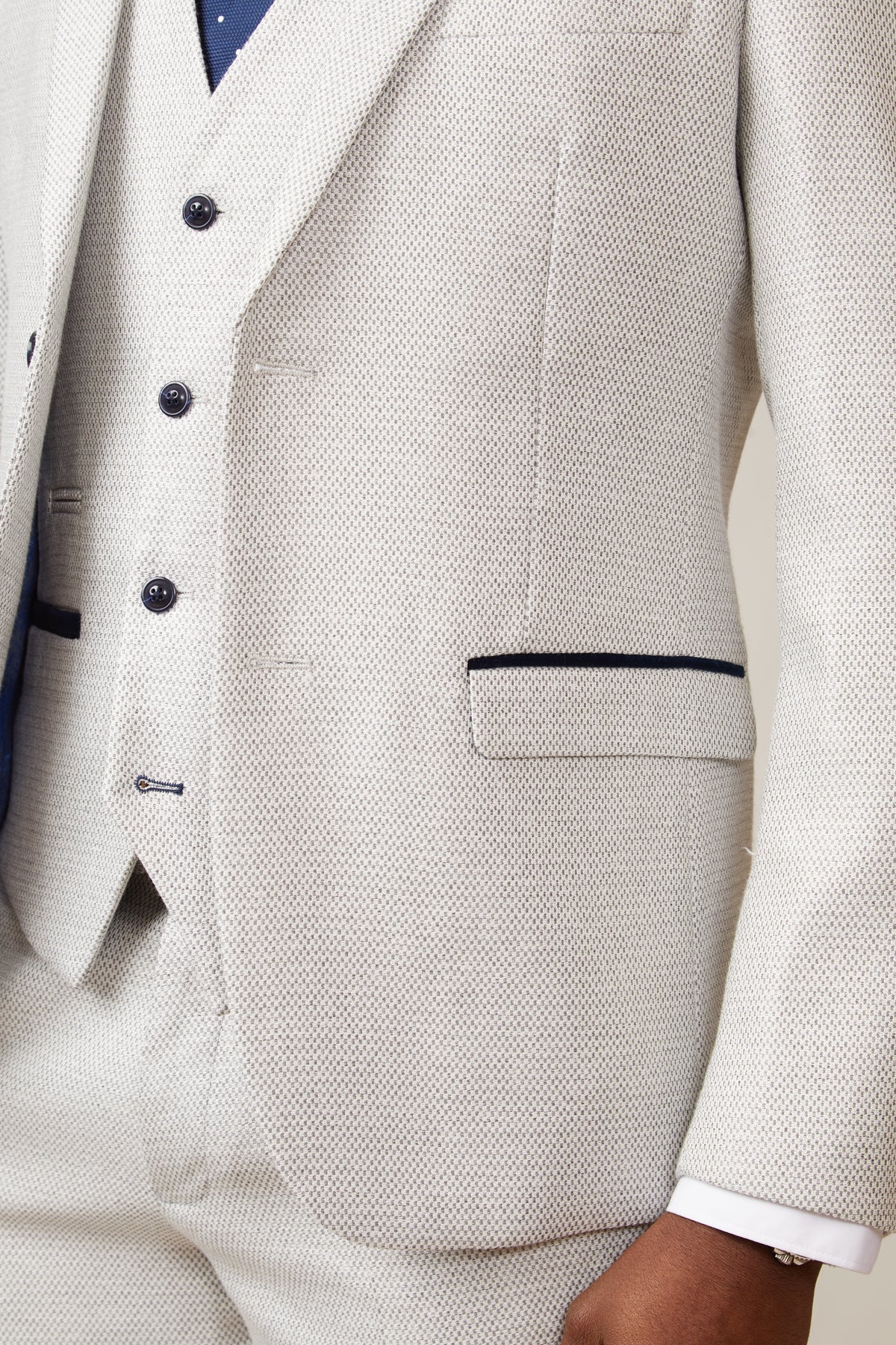 Close-up of a person wearing the marcdarcy RONALD - Stone Tailored Blazer with dark buttons, navy pocket trim, white shirt, and navy dot tie. Only the torso and arm are visible. The blazer features paisley print lining.