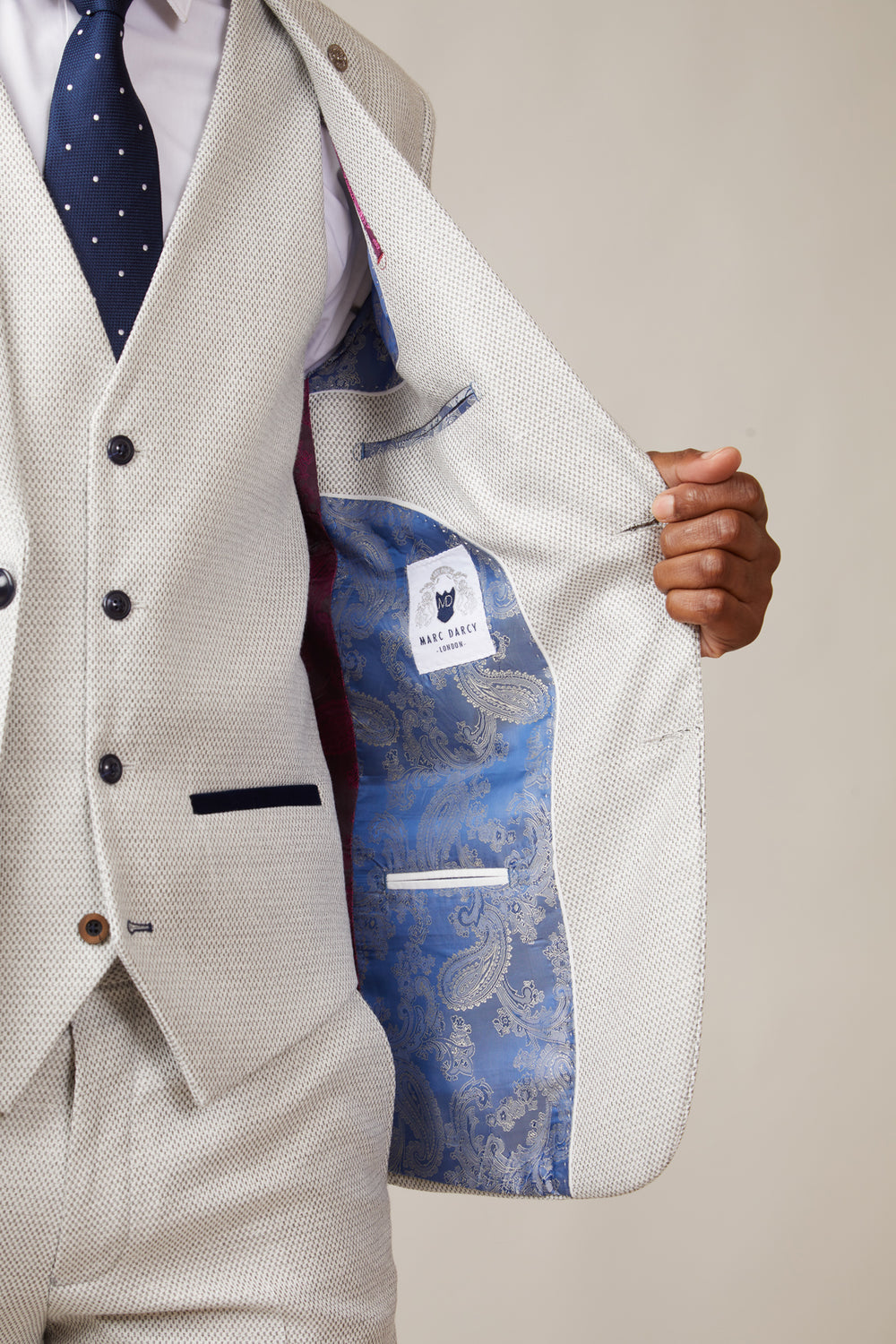 A man wears the marcdarcy RONALD Stone Tailored Blazer open, showing its bright blue paisley lining and brand label, paired with a dark blue polka dot tie and vest for a sharp slim fit look.