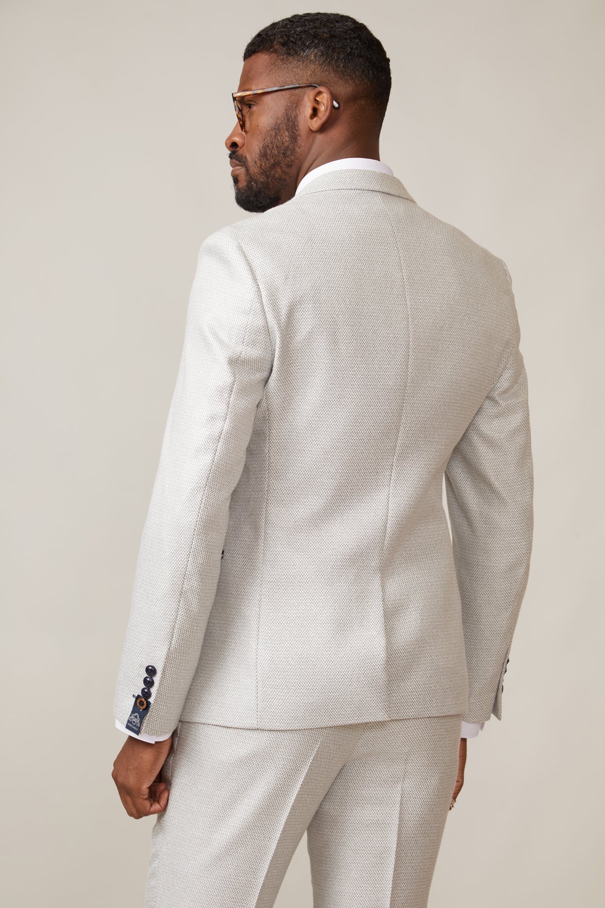 A man stands with his back to the camera, head turned left, wearing the marcdarcy RONALD - Stone Tailored Blazer with dark sleeve buttons and paisley lining. The background is plain and neutral.