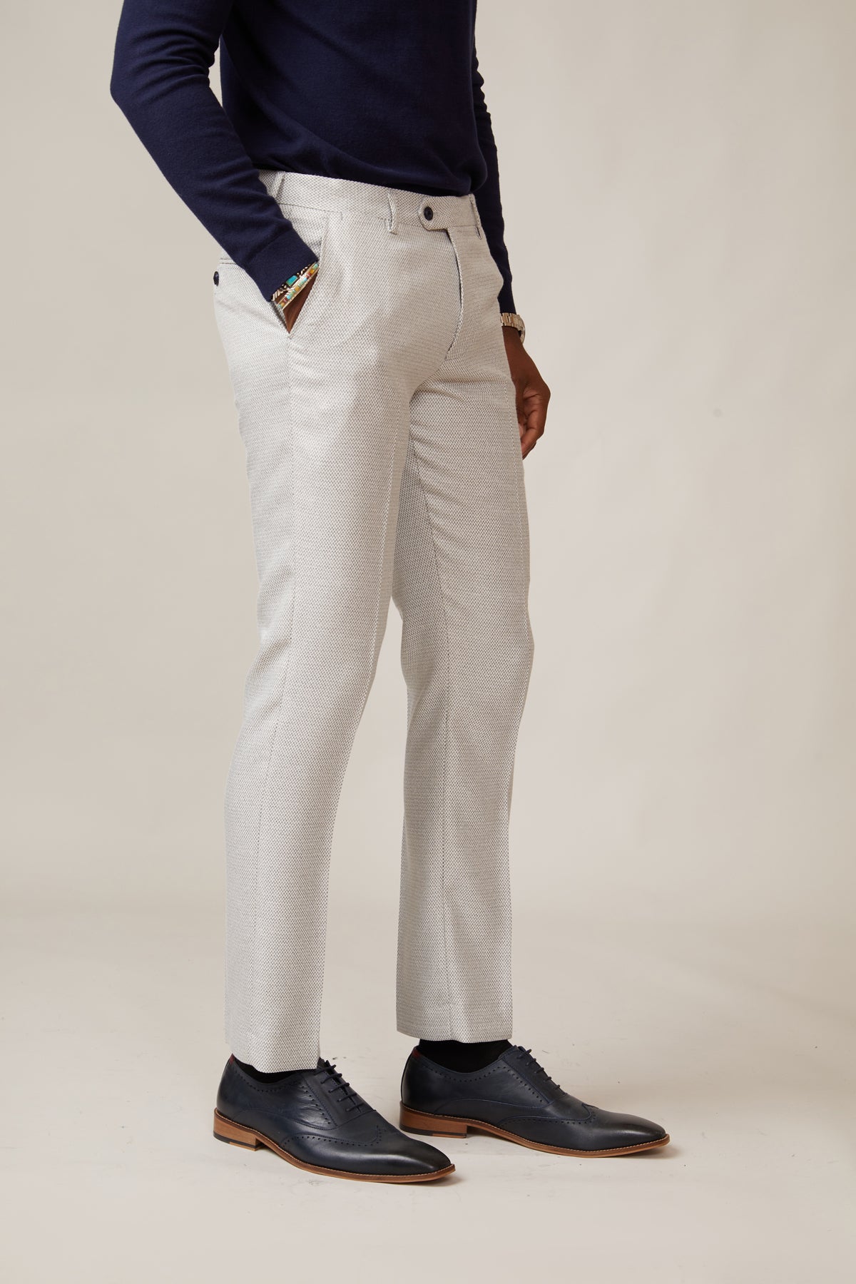 Wearing marcdarcy RONALD - Stone Tailored Trousers, a dark navy long-sleeve top, and black dress shoes, a person stands with hands in pockets against a plain light background. The head is not visible.