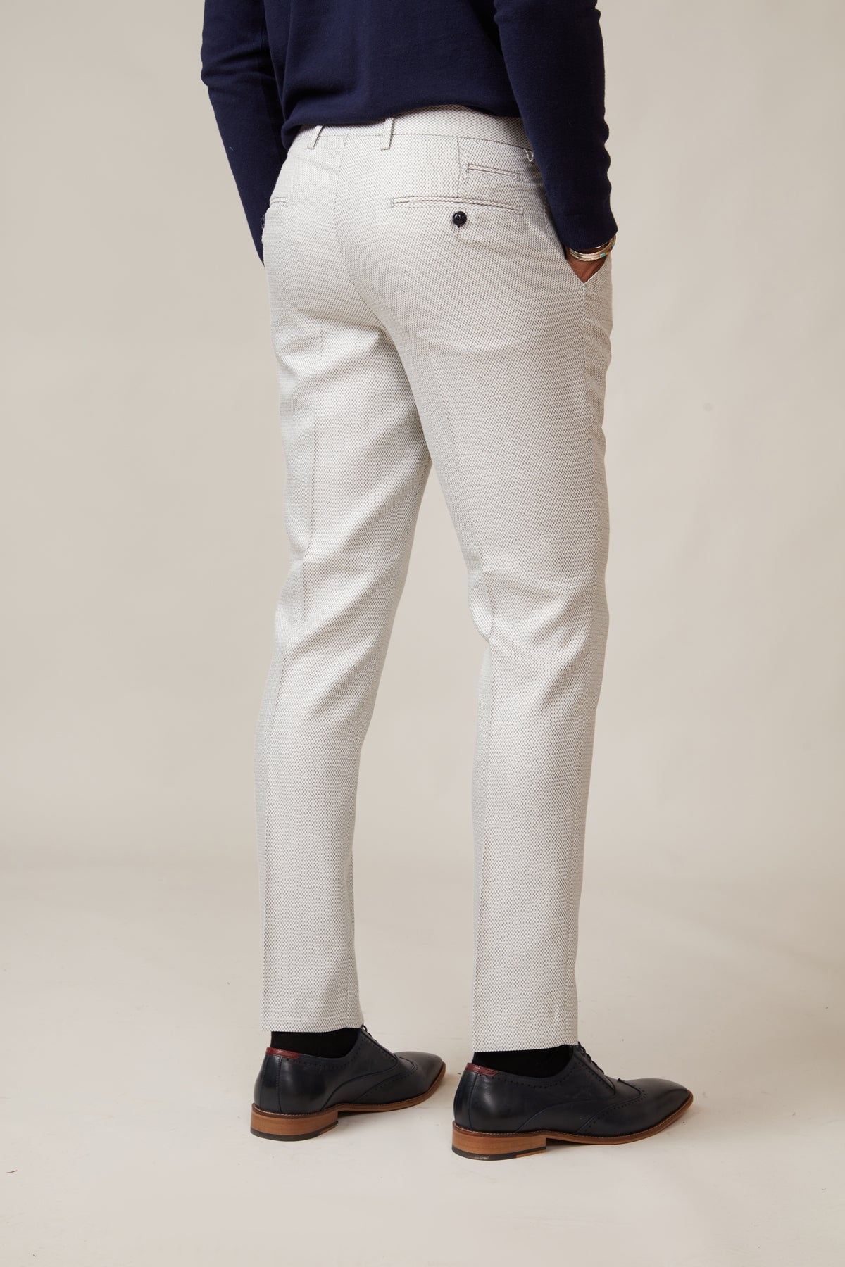 A person in marcdarcy's RONALD Stone Tailored Trousers, a dark navy sweater, and black dress shoes stands with their back to the camera against a plain light background.