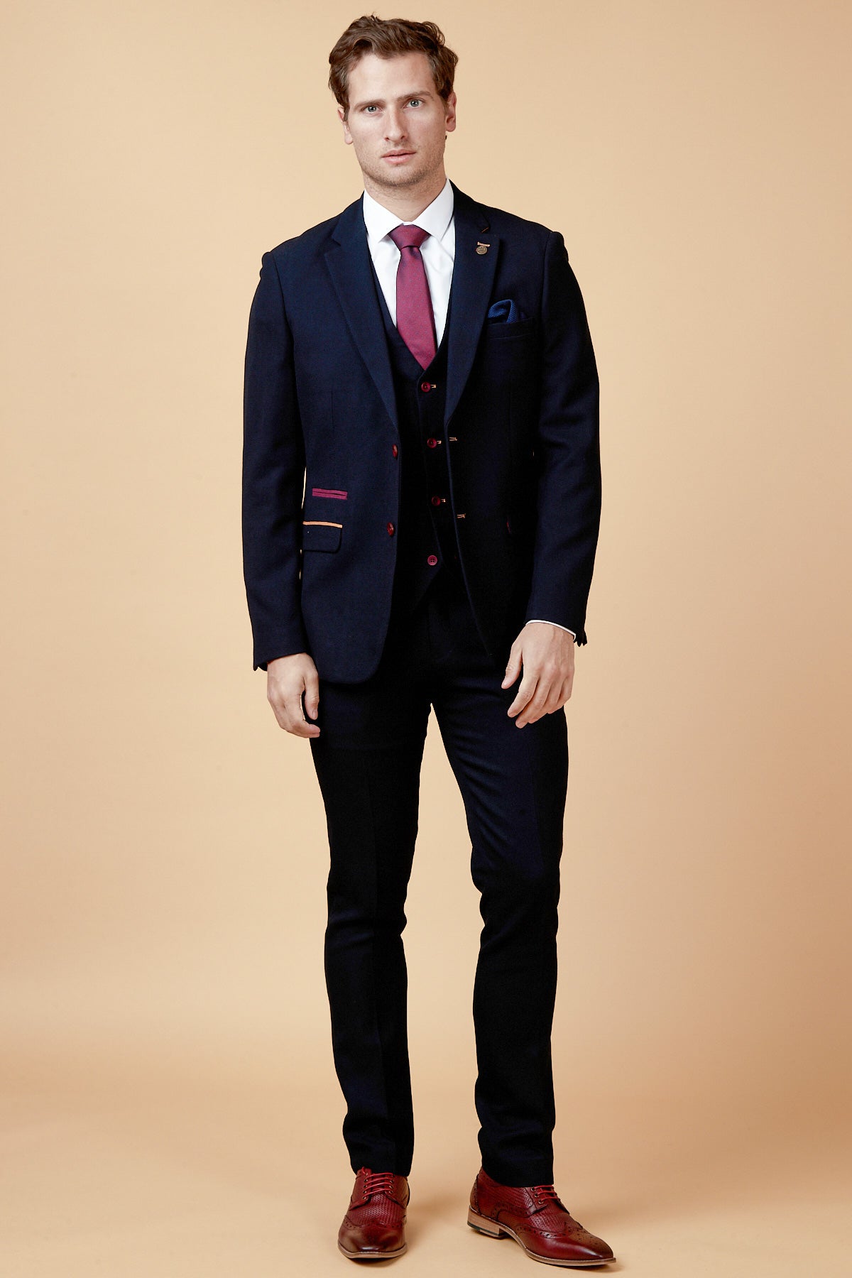 A man with short hair and a neutral expression stands against a beige background wearing the marcdarcy JD4 - Navy Contrast Trim Three Piece Suit, paired with a white shirt, maroon tie, and brown shoes—ideal attire for a stylish wedding guest.