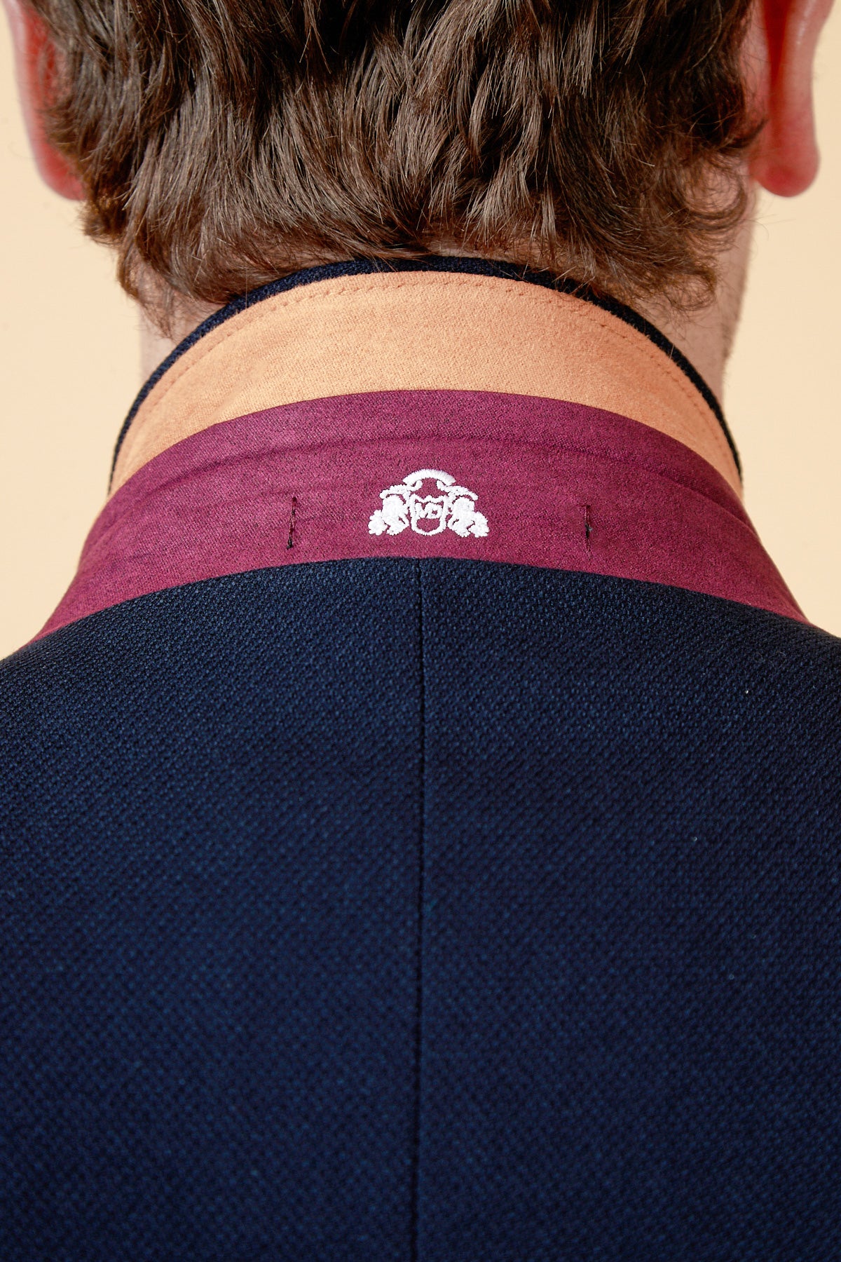 A close-up of the back of a person’s neck and shoulders highlights the marcdarcy JD4 - Navy Contrast Trim Two Piece Suit, featuring a burgundy and tan collar with a white embroidered crest and dark blue paisley-lined jacket. The persons hair is brown and wavy.
