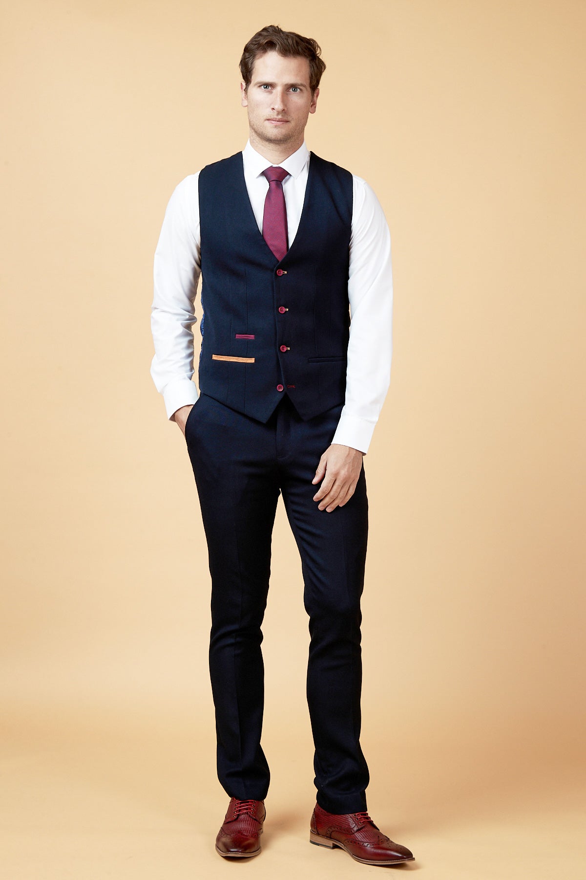 A man wears the marcdarcy JD4 - Navy Contrast Trim Waistcoat with a white shirt, burgundy tie, navy pants, and burgundy shoes, standing against a tan background.