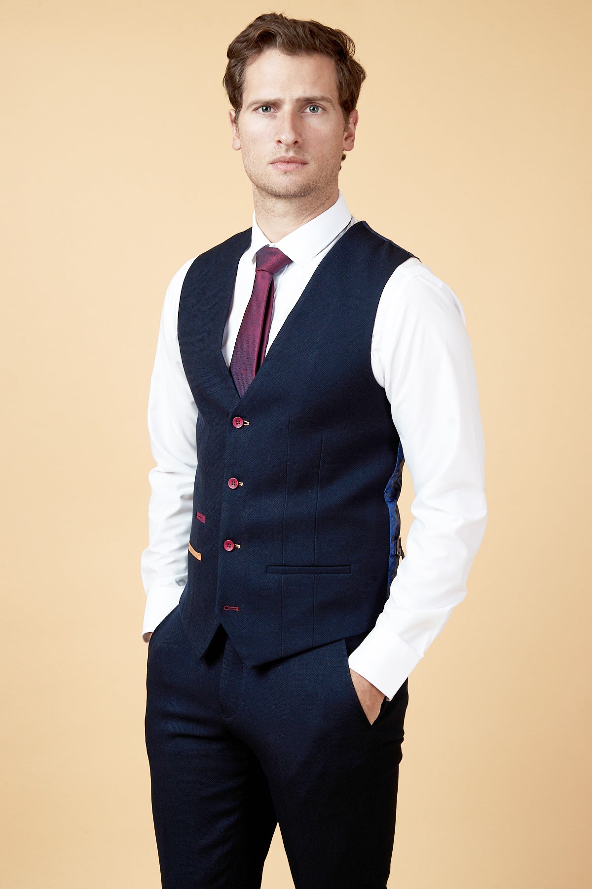 A man in a marcdarcy JD4 Navy Contrast Trim Waistcoat, white dress shirt, matching trousers, and a red tie stands with his hands in his pockets against a plain beige background.