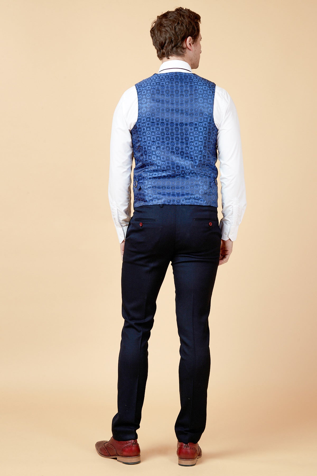 Man in a white dress shirt, navy JD4 - Navy Contrast Trim Waistcoat by marcdarcy with red buttons, and navy pants. Brown wingtip shoes complete his look against a cream background—perfect style for a wedding guest.