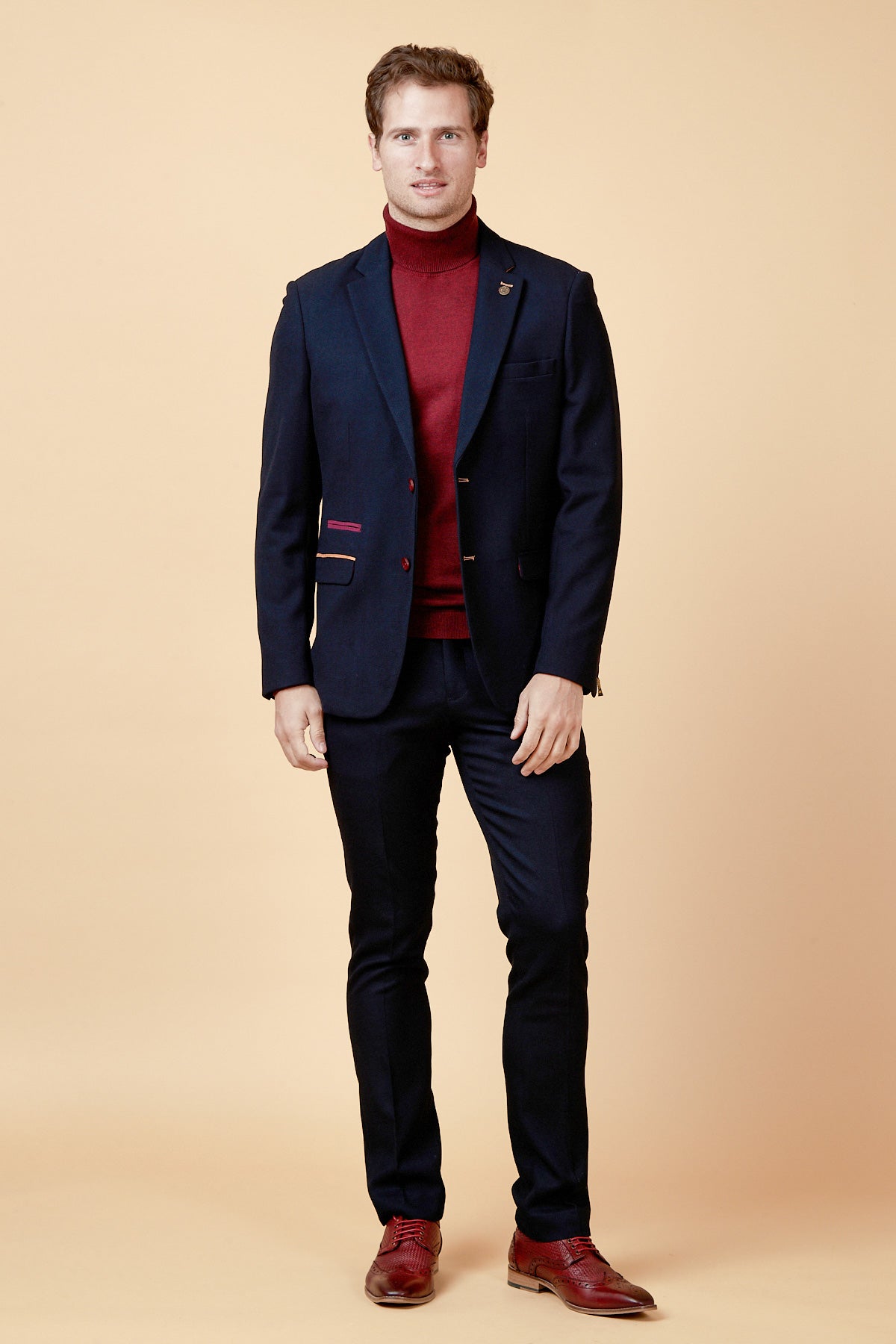 A man faces forward against a beige background, wearing the marcdarcy JD4 - Navy Contrast Trim Two Piece Suit with paisley lining, paired with a red turtleneck and red dress shoes—a standout look for a stylish wedding guest.