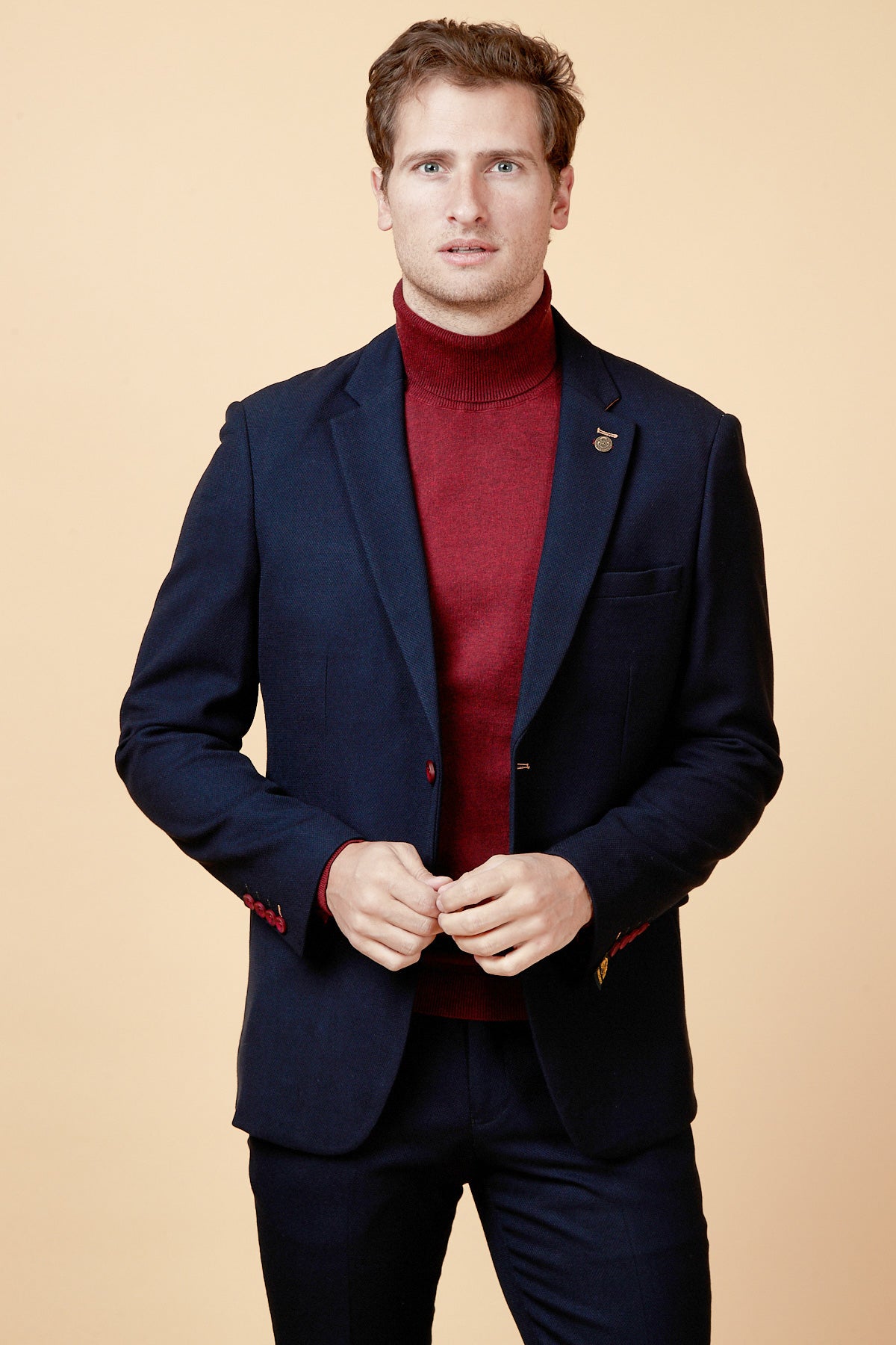A man models the marcdarcy JD4 - Navy Contrast Trim Blazer over a red turtleneck, standing against a beige background and looking at the camera with his hands clasped, highlighting sophisticated workwear style.
