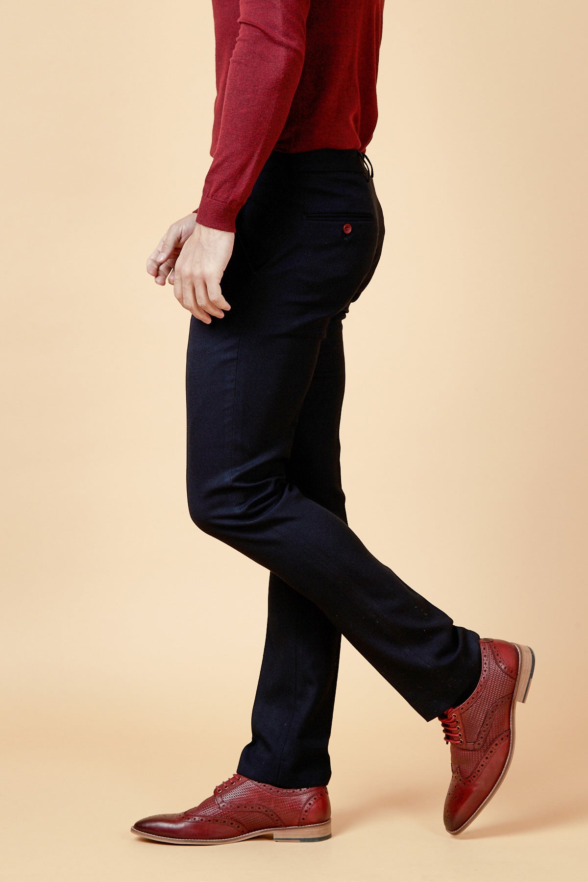 A man wearing marcdarcy JD4 Navy Flat Front Trousers, paired with a maroon sweater and red wingtip shoes, walks against a tan background.