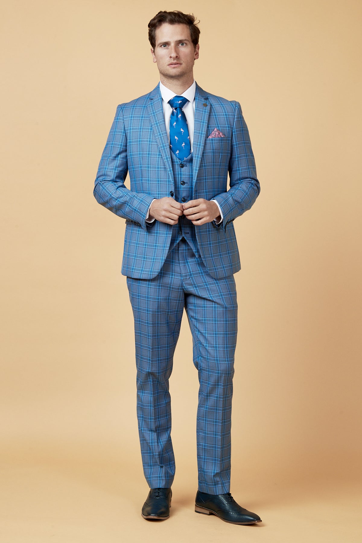 A man stands against a beige background in a marcdarcy JOSE Sky Blue Check Three Piece Suit with matching vest, white shirt, patterned blue tie, and dark dress shoes. He looks forward, adjusting his slim fit suit jacket.