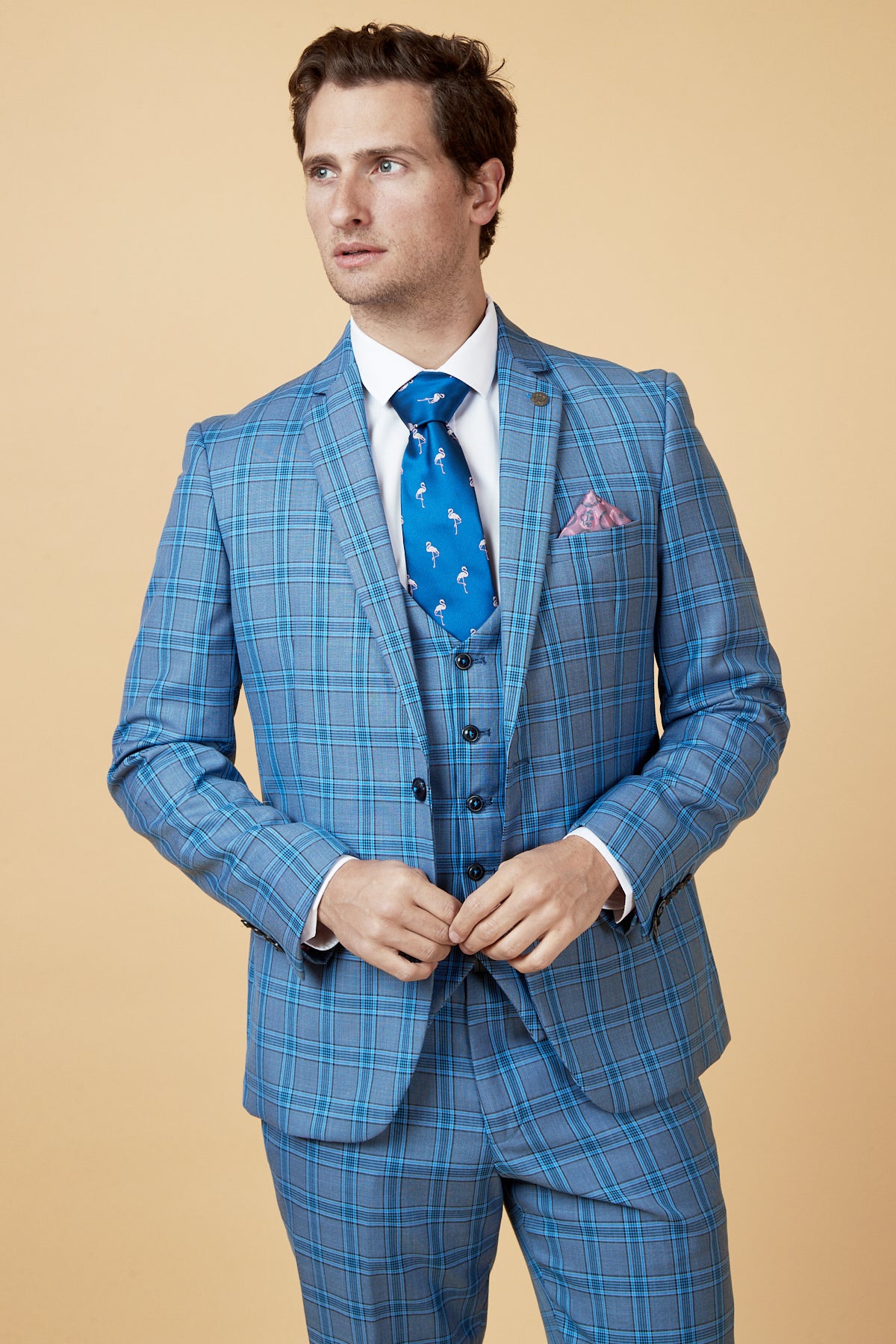 A man wearing the marcdarcy JOSE Sky Blue Check Three Piece Suit with a white shirt, blue patterned tie, and pink pocket square stands against a beige background, looking slightly to the side.