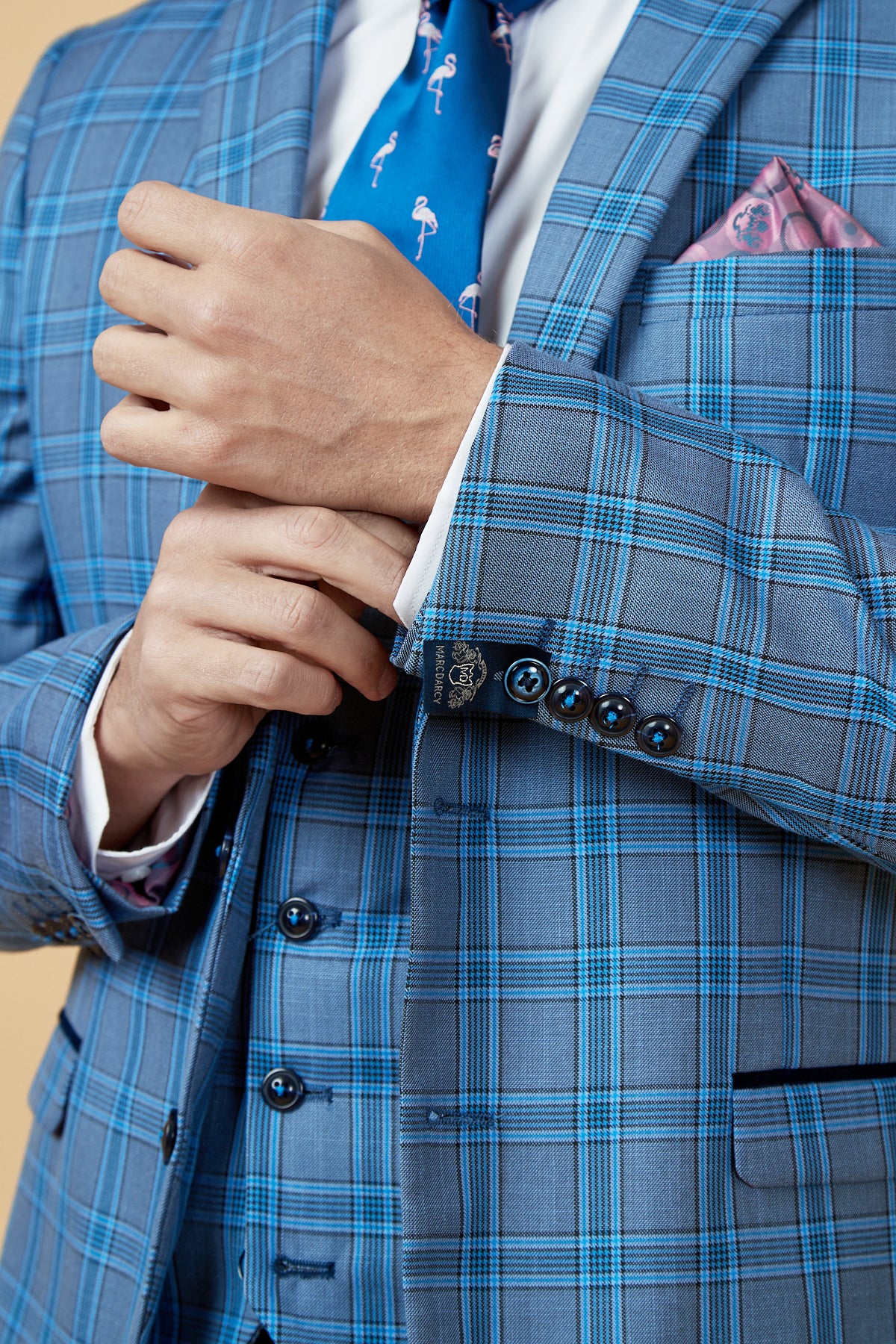 A man adjusts the cuff of his Marc Darcy JOSE Sky Blue Check Blazer, paired with a matching vest, blue flamingo tie, and pink pocket square. Only his torso and hands are visible.