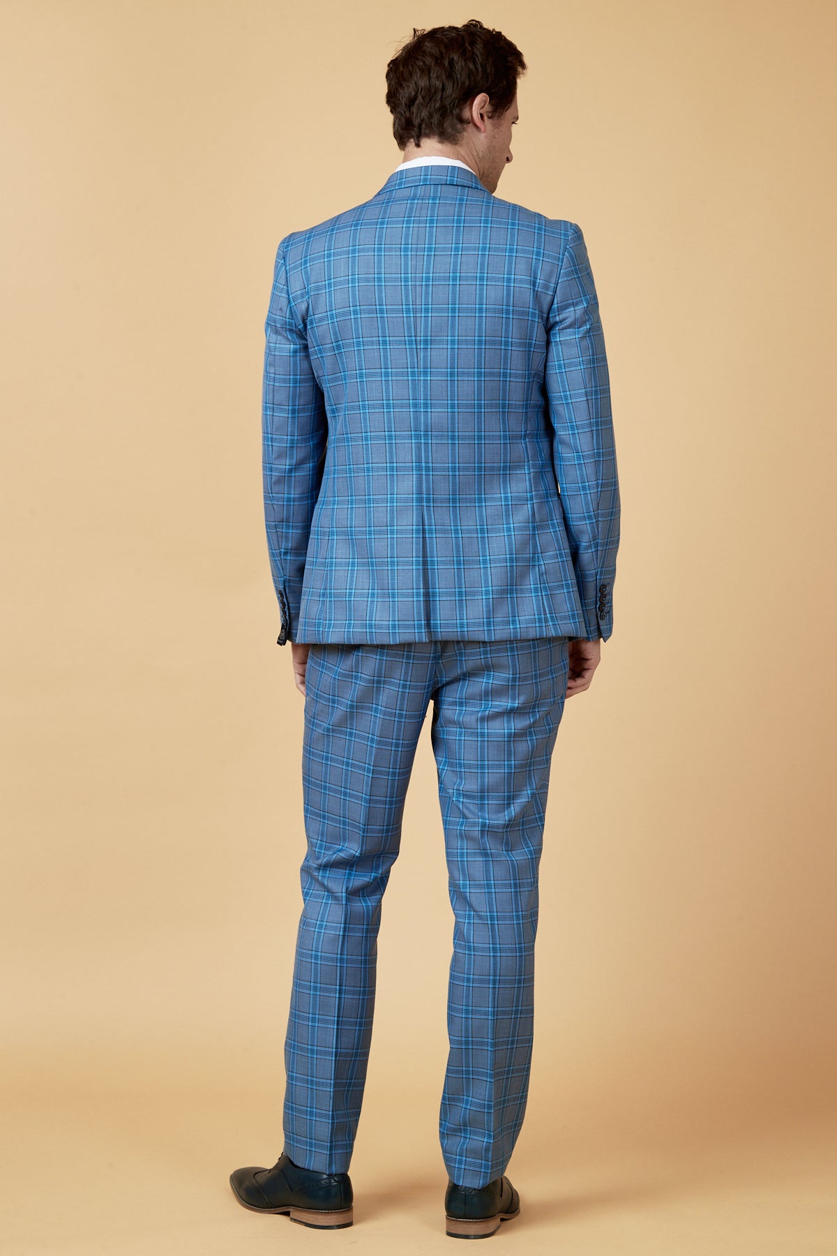 A man stands with his back to the camera, wearing a marcdarcy JOSE Sky Blue Check Three Piece Suit and a white shirt, with black shoes, against a plain beige background.