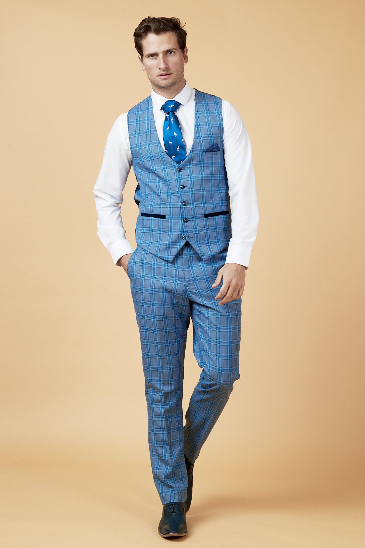 A man stands against a beige background wearing the marcdarcy JOSE Sky Blue Check Three Piece Suit, paired with a white dress shirt and a blue tie with white and navy designs. With one hand in his pocket, he looks confidently at the camera.