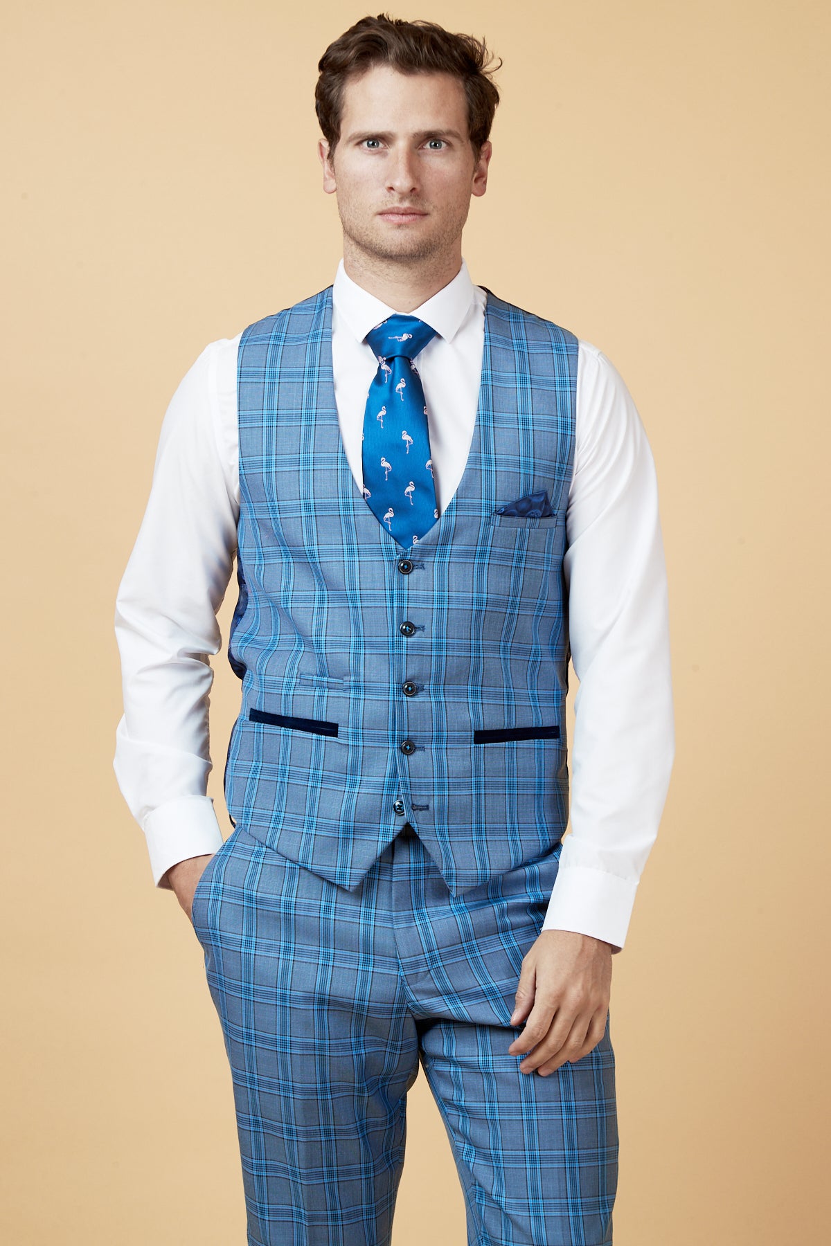 A man stands against a beige background wearing the marcdarcy JOSE Sky Blue Check Single Breasted Waistcoat with matching pants, a white dress shirt, and a patterned blue tie, one hand in his pocket as he looks at the camera.