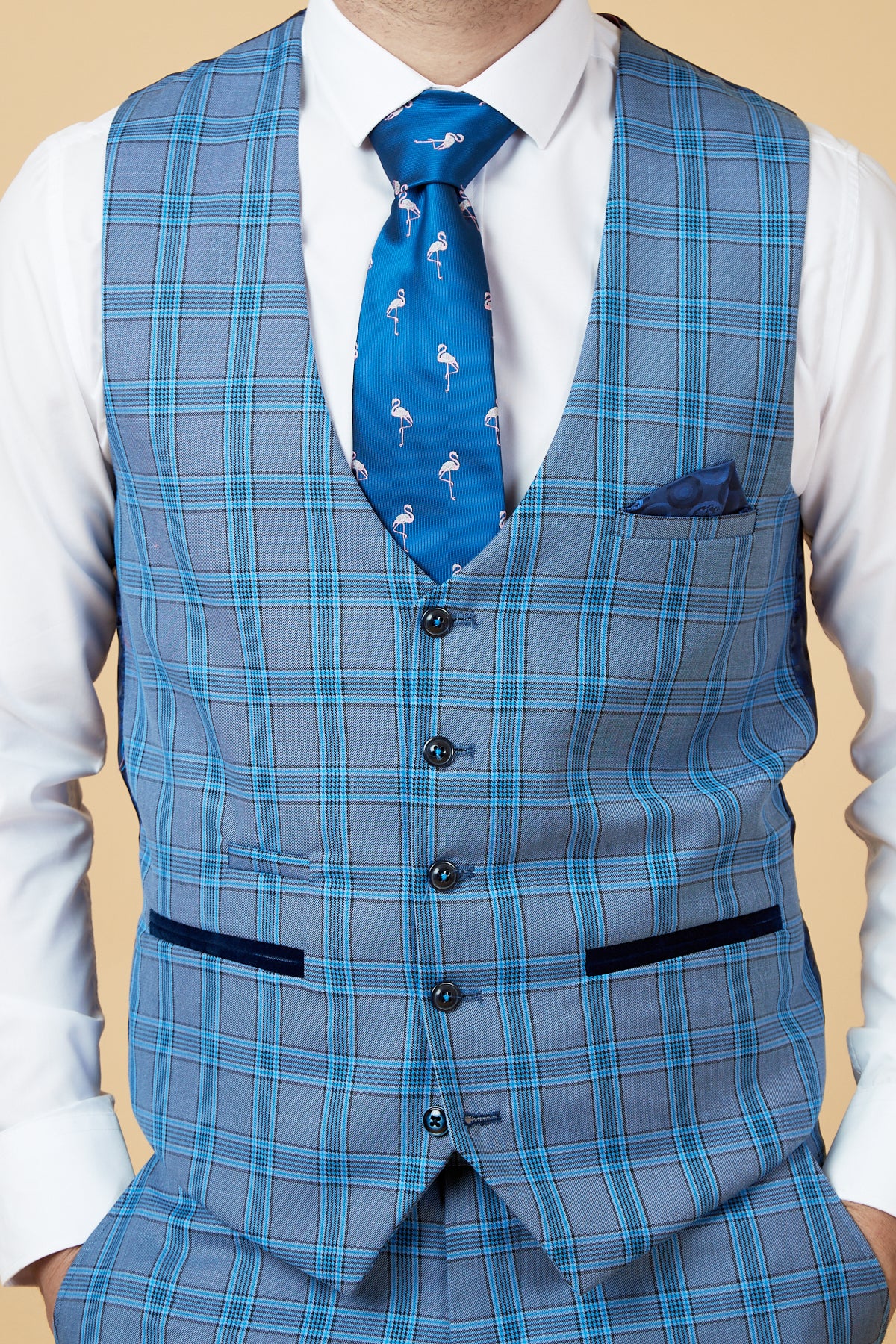 A man wears a marcdarcy JOSE Sky Blue Check Single Breasted Waistcoat, slim fit blue plaid three-piece suit, white dress shirt, blue flamingo-pattern tie, and pocket square, standing hands-in-pockets against a beige background.