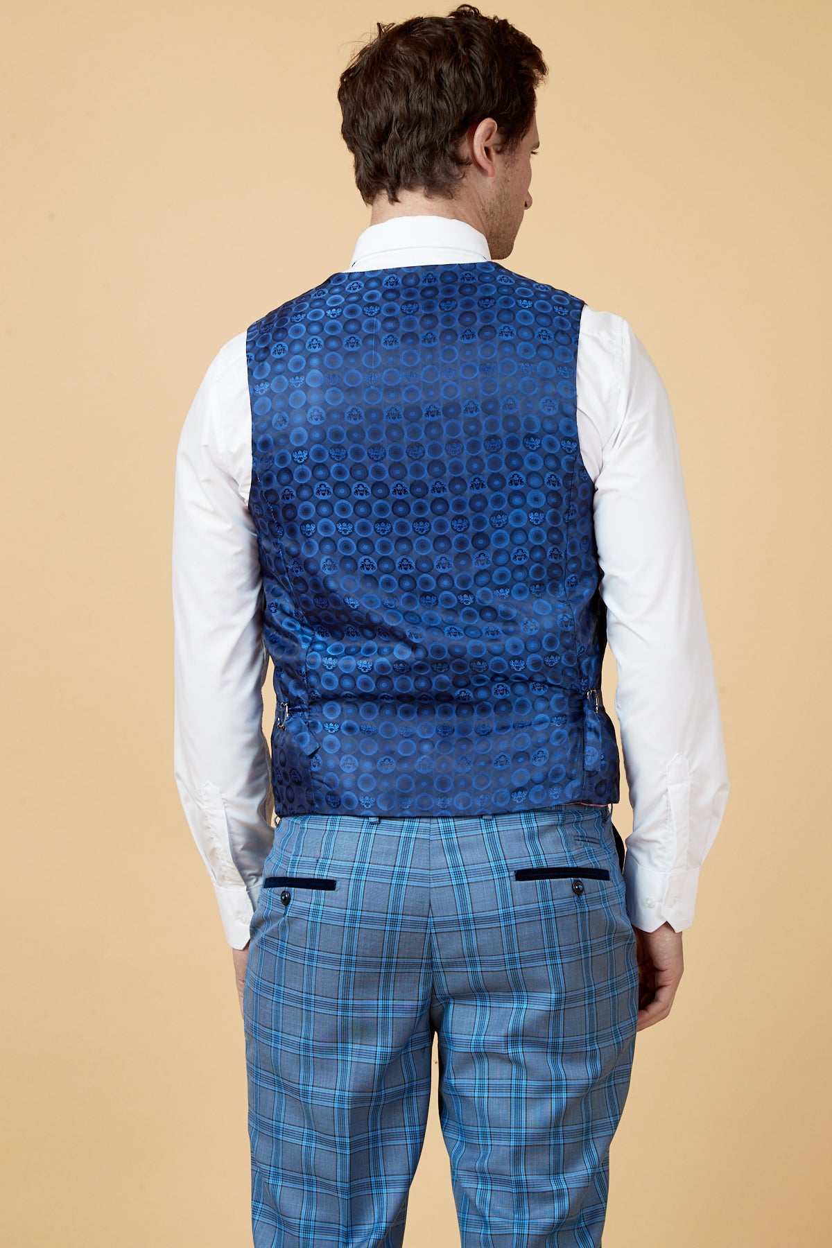 A man stands with his back to the camera, wearing a white shirt and the marcdarcy JOSE Sky Blue Check Three Piece Suit against a beige background.