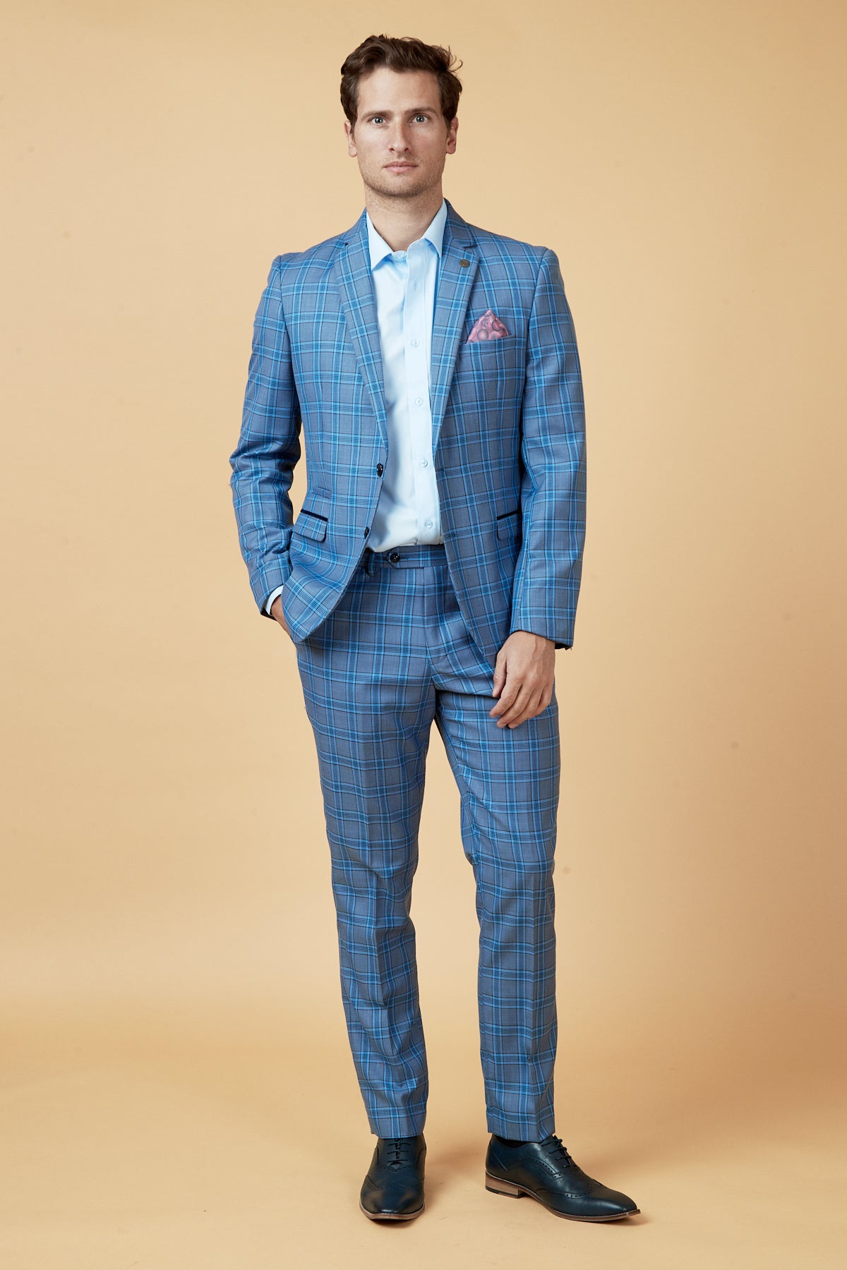 A man poses confidently against a beige background wearing the Marc Darcy JOSE - Sky Blue Check Blazer, a light blue dress shirt, dark shoes, and a pink pocket square, with one hand in his pocket.