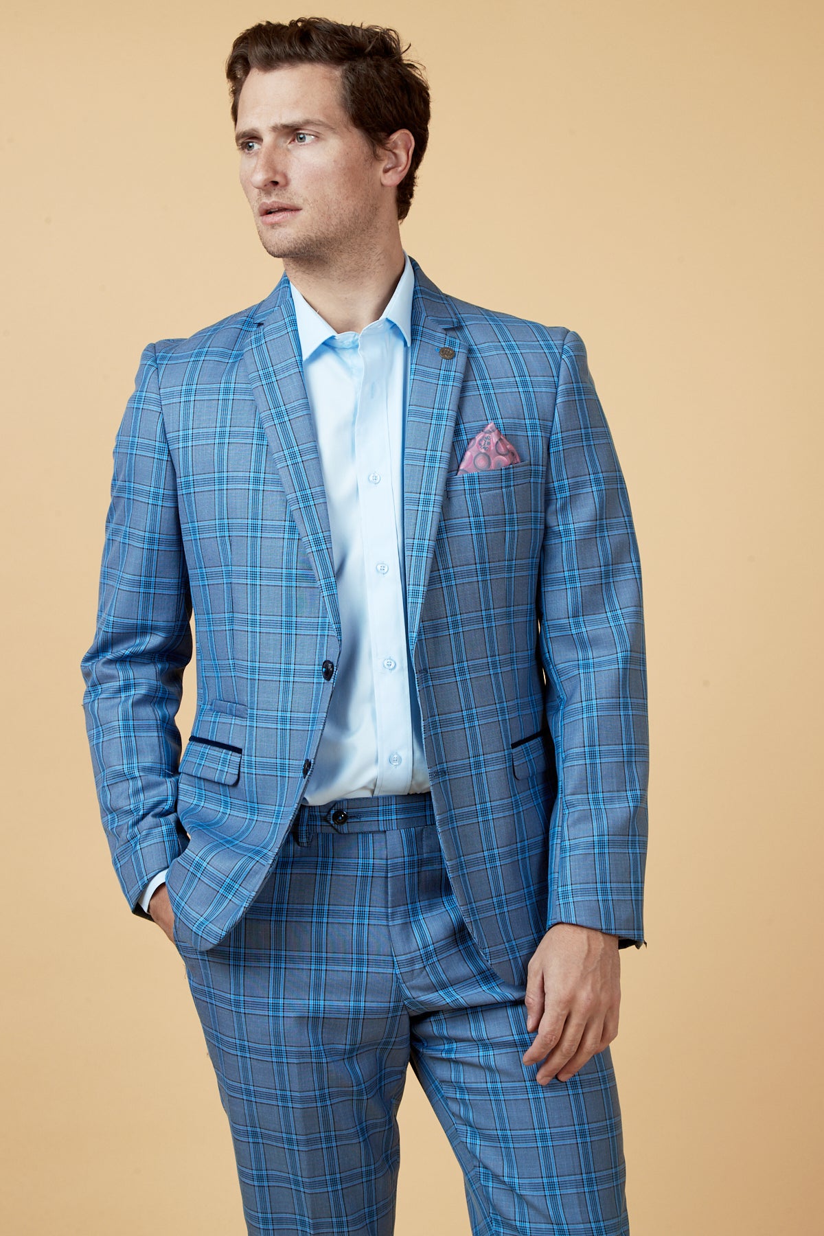 A man wears the Marc Darcy JOSE - Sky Blue Check Blazer with a light blue shirt and pink pocket square, standing against a beige background and looking to the side with one hand in his pocket.