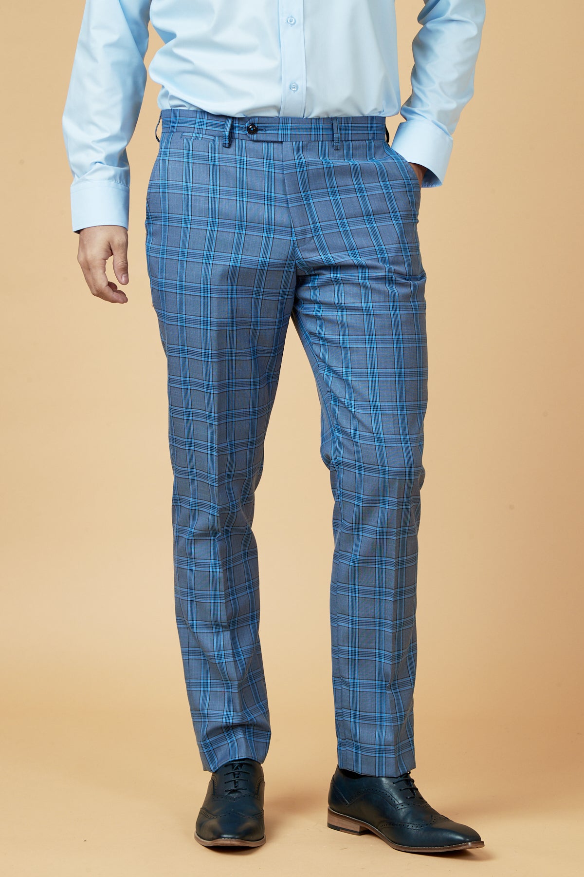 A man dressed in a marcdarcy JOSE sky blue check three piece suit, paired with a light blue shirt and dark blue shoes, stands against a beige background with his hands in his pockets.