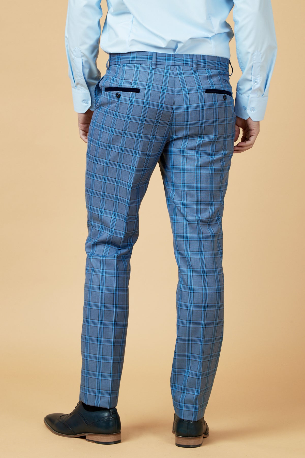 A man faces away, wearing marcdarcy’s JOSE Sky Blue Check Trousers with a light blue shirt and black dress shoes, set against a beige background.