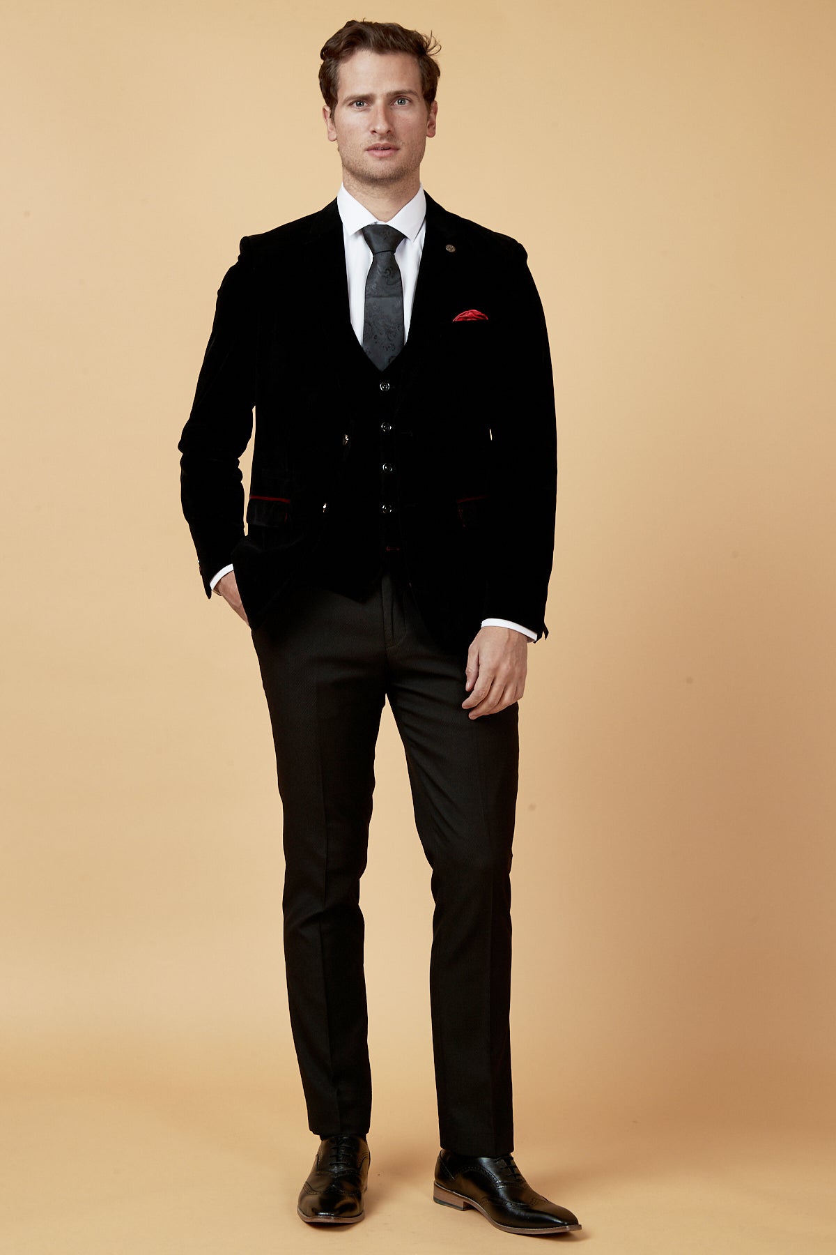 A man stands against a beige background in the marcdarcy HUDSON black velvet blazer, white shirt, black tie, black dress shoes, and a red pocket square. One hand is in his pocket as he looks straight ahead.