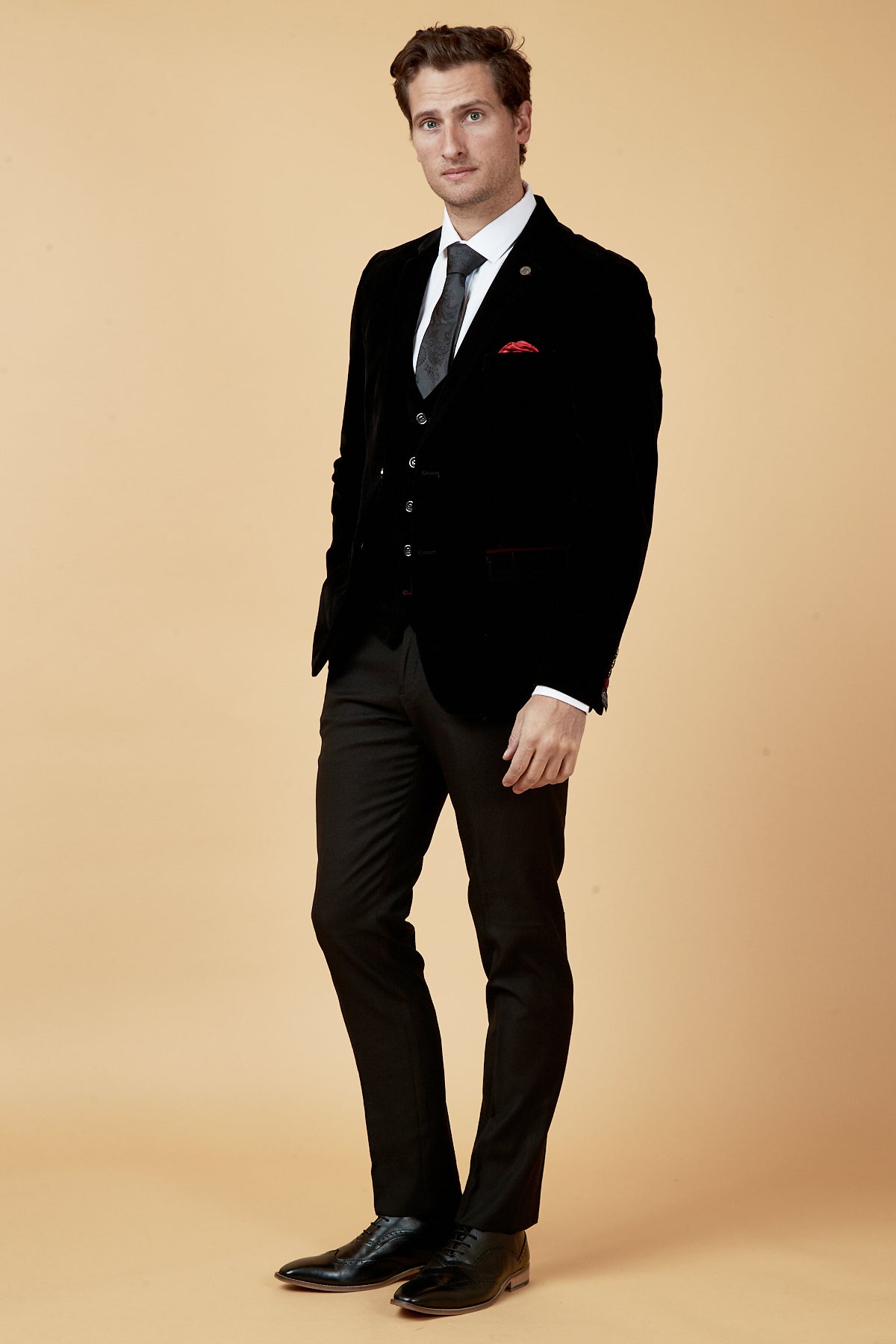 Wearing the marcdarcy HUDSON Black Velvet Blazer with a white shirt, black tie, and red pocket square, a man stands against a light beige background, gazing at the camera with a subtle tilt to his posture.