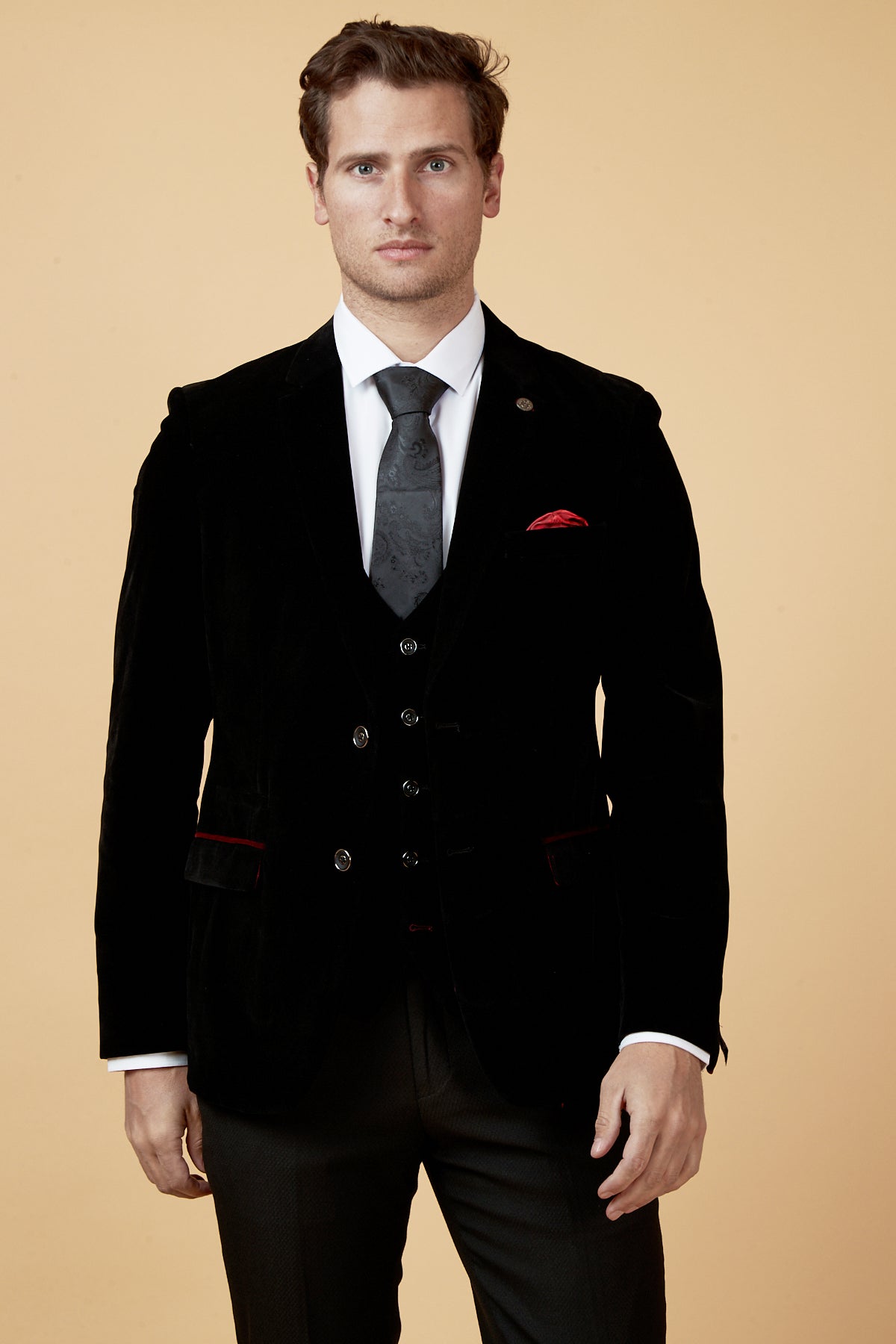 A man faces the camera against a beige backdrop, wearing marcdarcy's HUDSON Black Velvet Blazer with a white dress shirt, black patterned tie, dark trousers, and a red pocket square accenting his jacket.