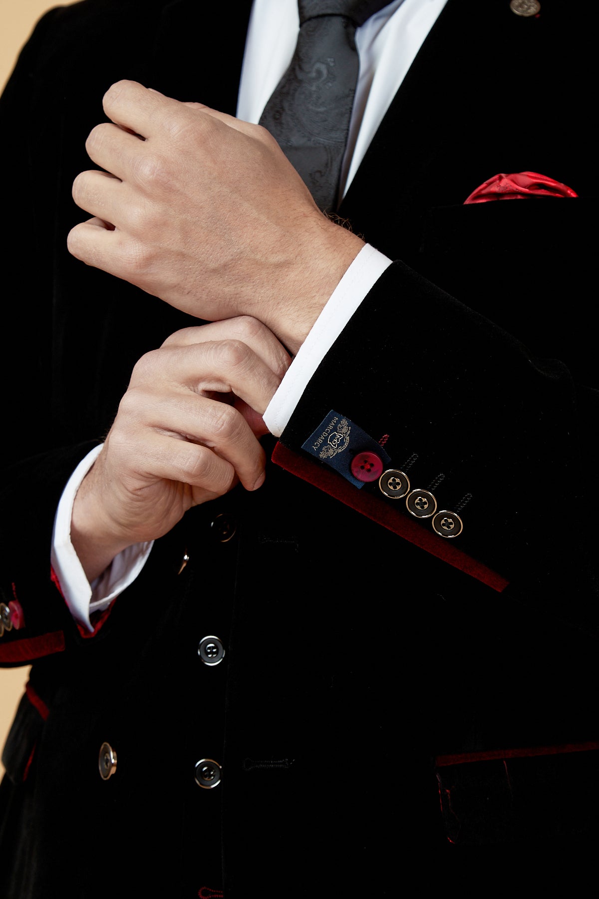 A man wearing the marcdarcy HUDSON Black Velvet Blazer with red accents adjusts his cuff. He pairs it with a white shirt, dark vest, patterned tie, and red pocket square. Only his torso and hands are shown.