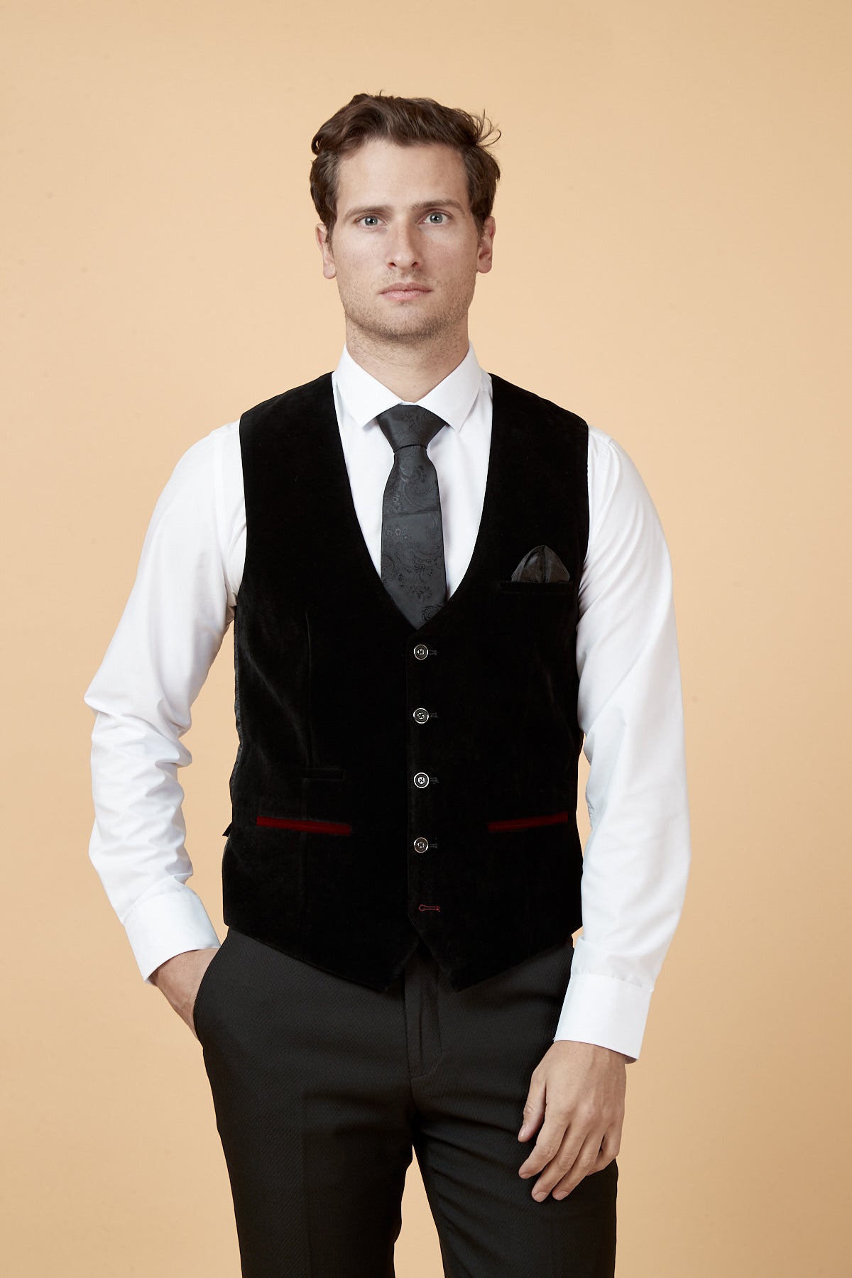 A man stands against a beige background, wearing the marcdarcy HUDSON Black Velvet Jacquard Waistcoat over a white dress shirt and black tie, with one hand in his pocket and a neutral expression.