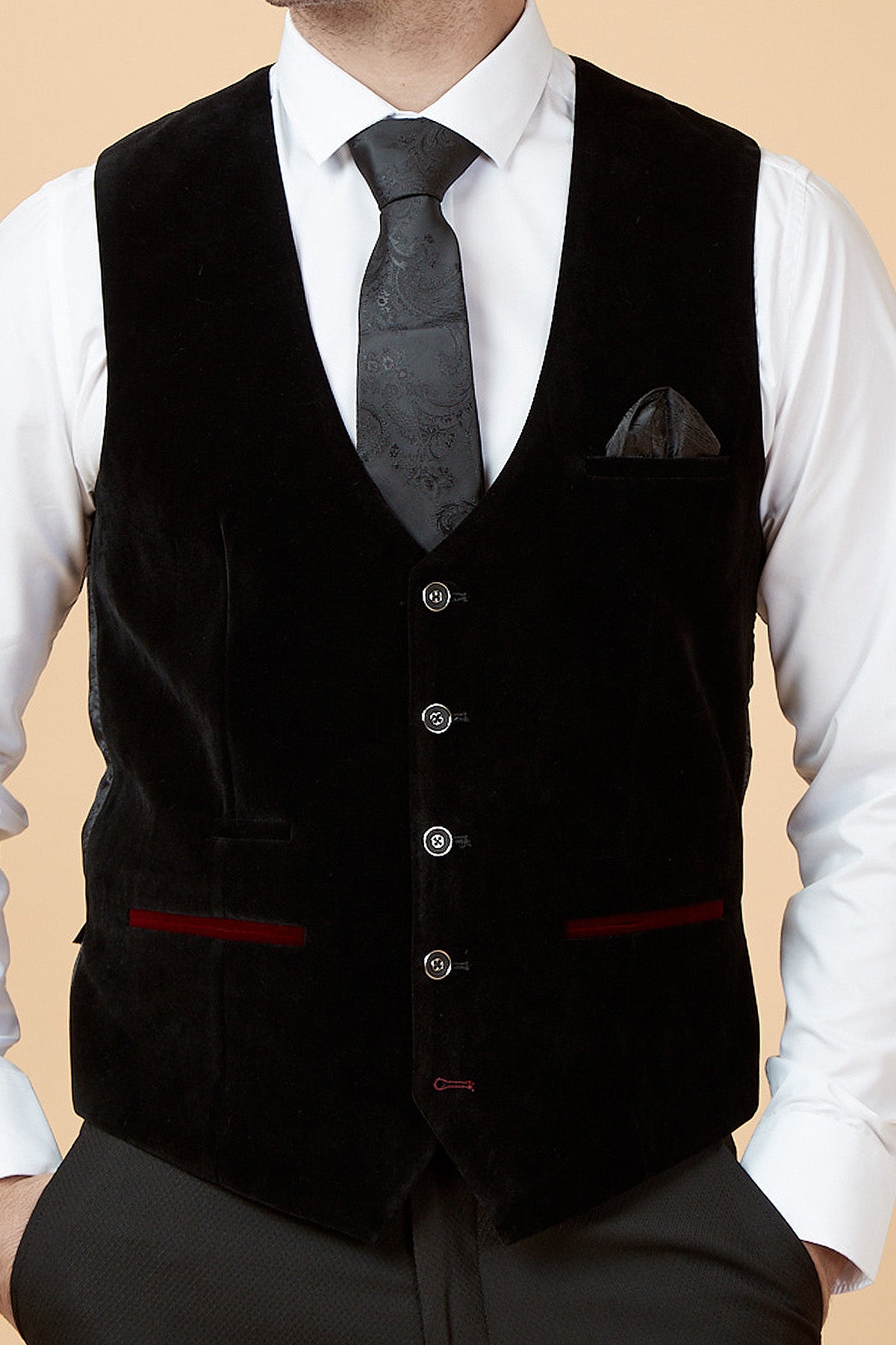A man wearing a white shirt, black patterned tie, dark pocket square, and the marcdarcy HUDSON Black Velvet Jacquard Waistcoat with tailored fit, buttons, and maroon-trimmed pockets stands with his hands in his pants pockets.