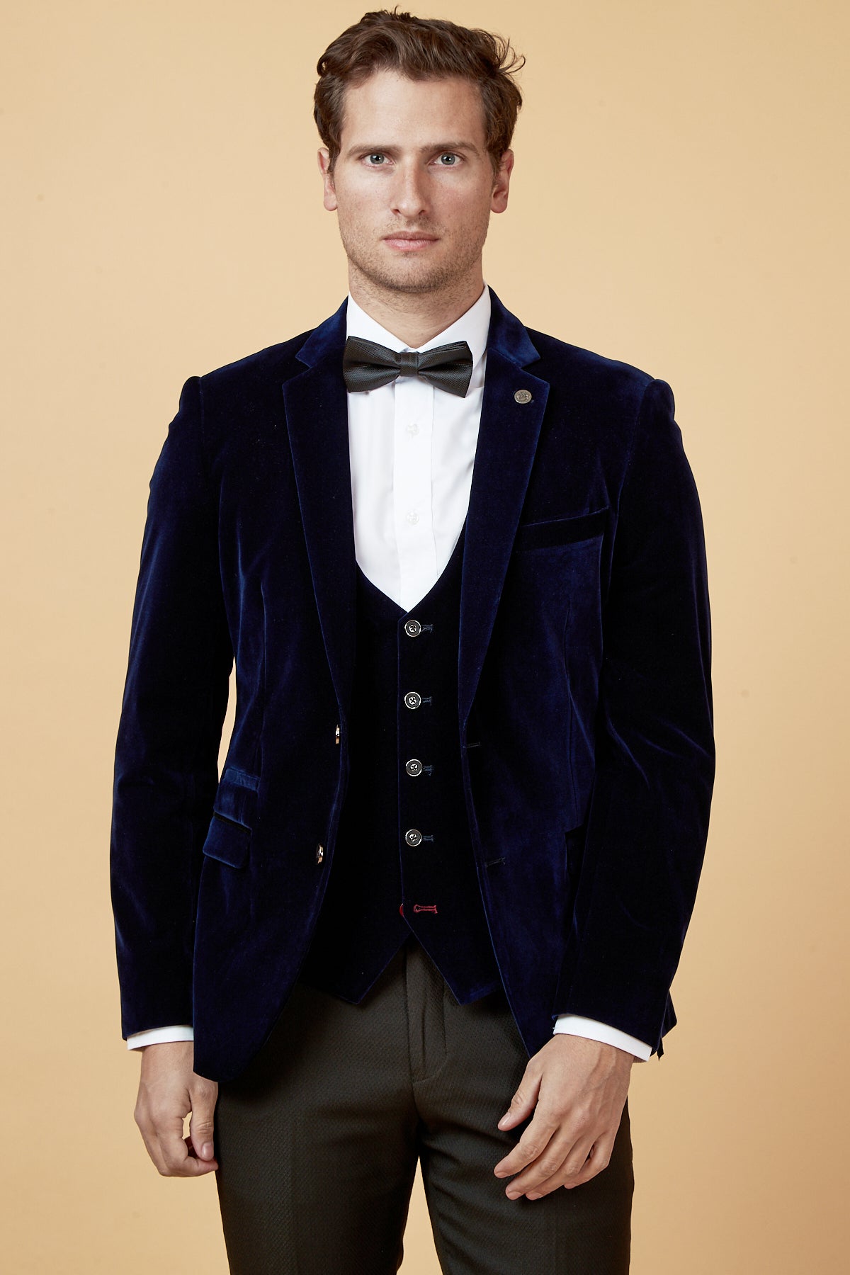 A man wearing the marcdarcy HUDSON Navy Velvet Blazer, a black waistcoat, white dress shirt, black bow tie, and dark trousers stands against a plain beige background, looking directly at the camera.