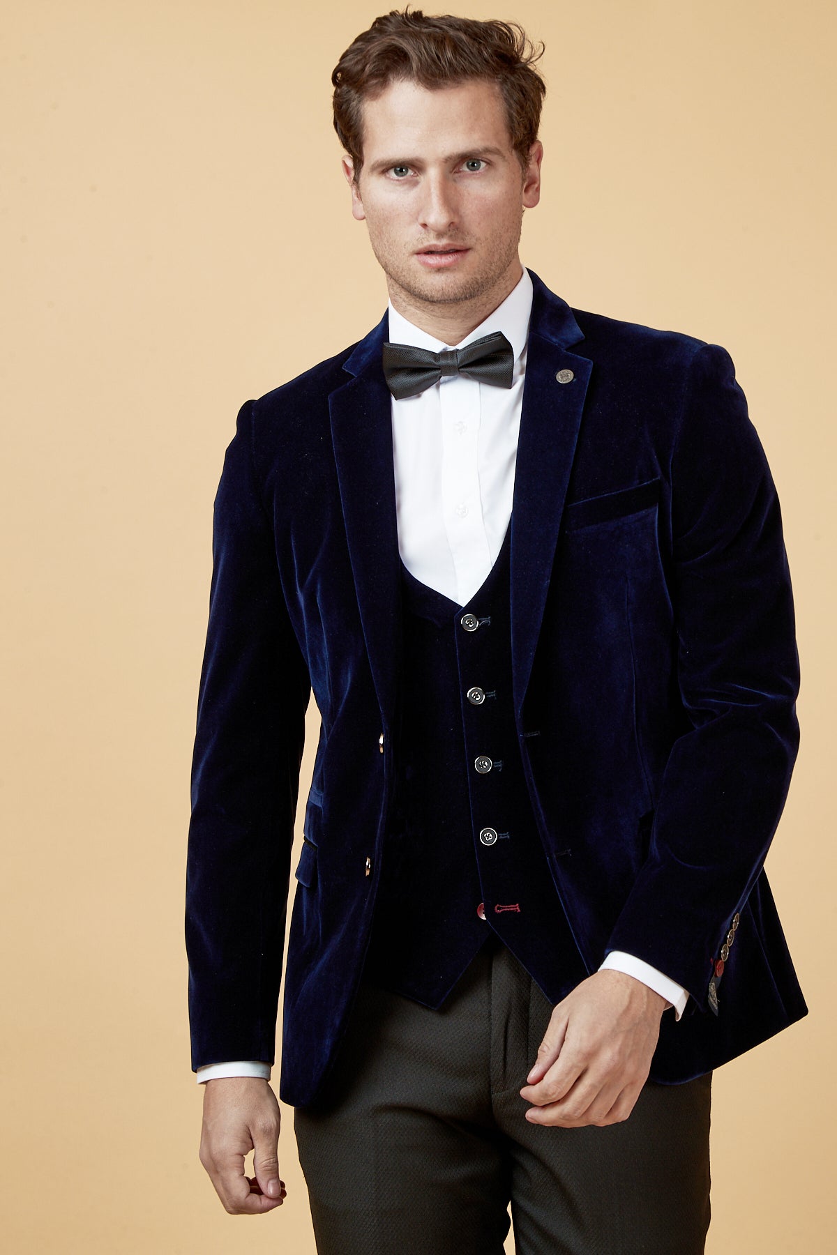A man wears the marcdarcy HUDSON Navy Velvet Blazer with a white shirt and black bowtie, standing against a beige background.