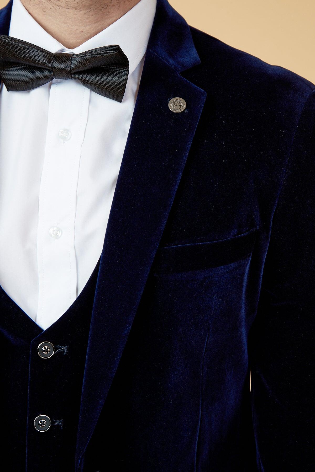 A person wears the marcdarcy HUDSON Navy Velvet Blazer with a pin on the lapel, paired with a white dress shirt and black bow tie, shown from chest to neck against a beige background.