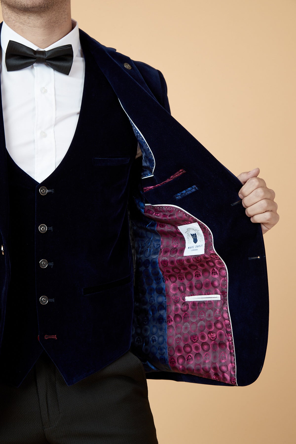 A man in a white shirt, black bow tie, and dark suit opens his marcdarcy HUDSON Navy Velvet Blazer to reveal its vibrant inner lining with red and blue patterns, multiple pockets, and a designer label.