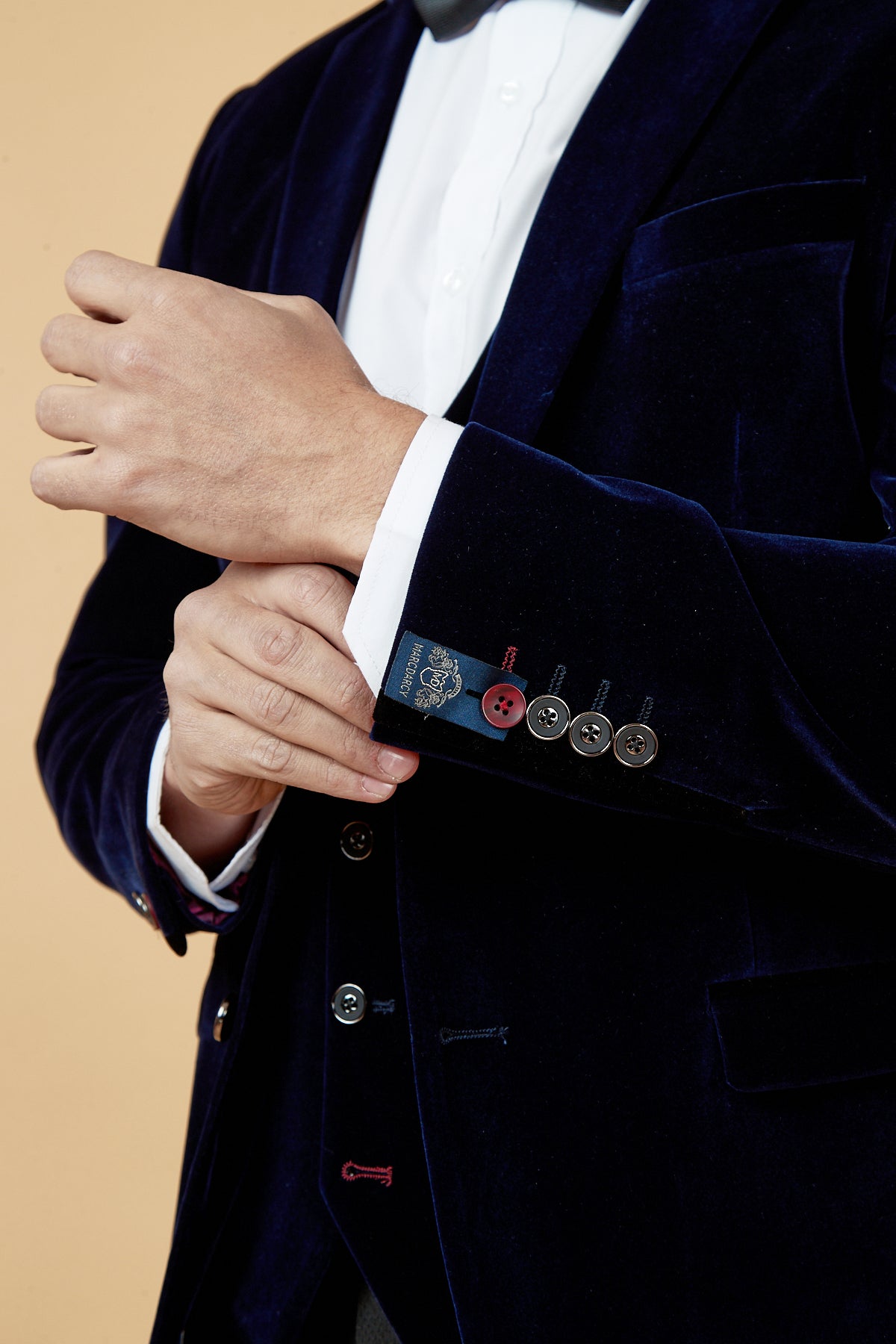 A man in the marcdarcy HUDSON Navy Velvet Blazer adjusts his shirt cuff, showcasing multicolored sleeve buttons and a visible inside label, set against a light beige background.