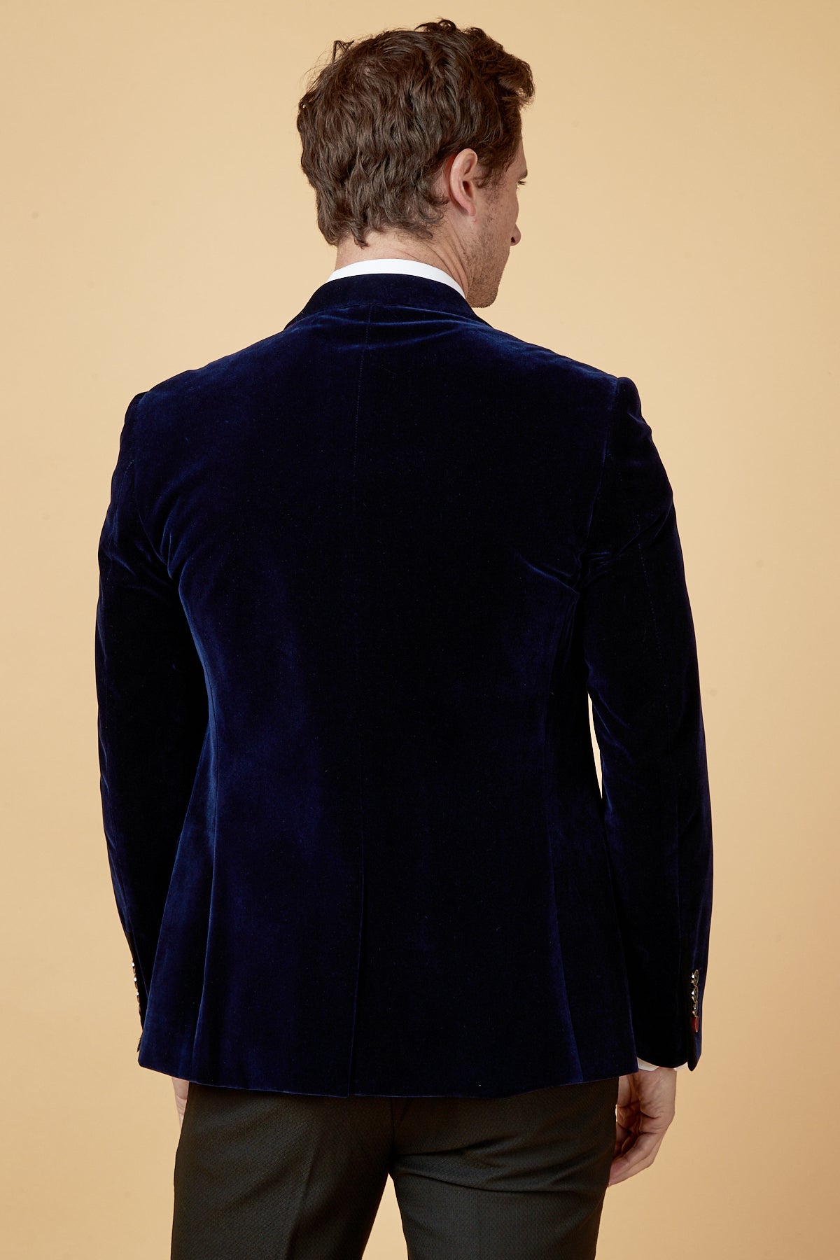 A man wears the marcdarcy HUDSON Navy Velvet Blazer over a white shirt and dark trousers, shown from the back against a tan background, highlighting the slim fit blazer’s sharp silhouette.