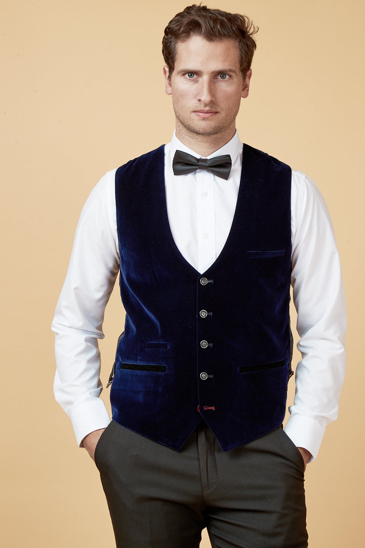 A man stands with his hands in his pockets against a beige background, wearing a white dress shirt, black bow tie, and the marcdarcy HUDSON - Navy Velvet Jacquard Waistcoat.