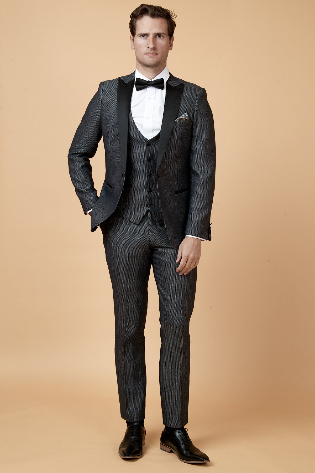 A man wearing the marcdarcy SPENCER Charcoal Grey Tux Lapel Three Piece Suit, paired with a white shirt, black vest, and black bow tie, stands with his hand in his pocket against a beige background.