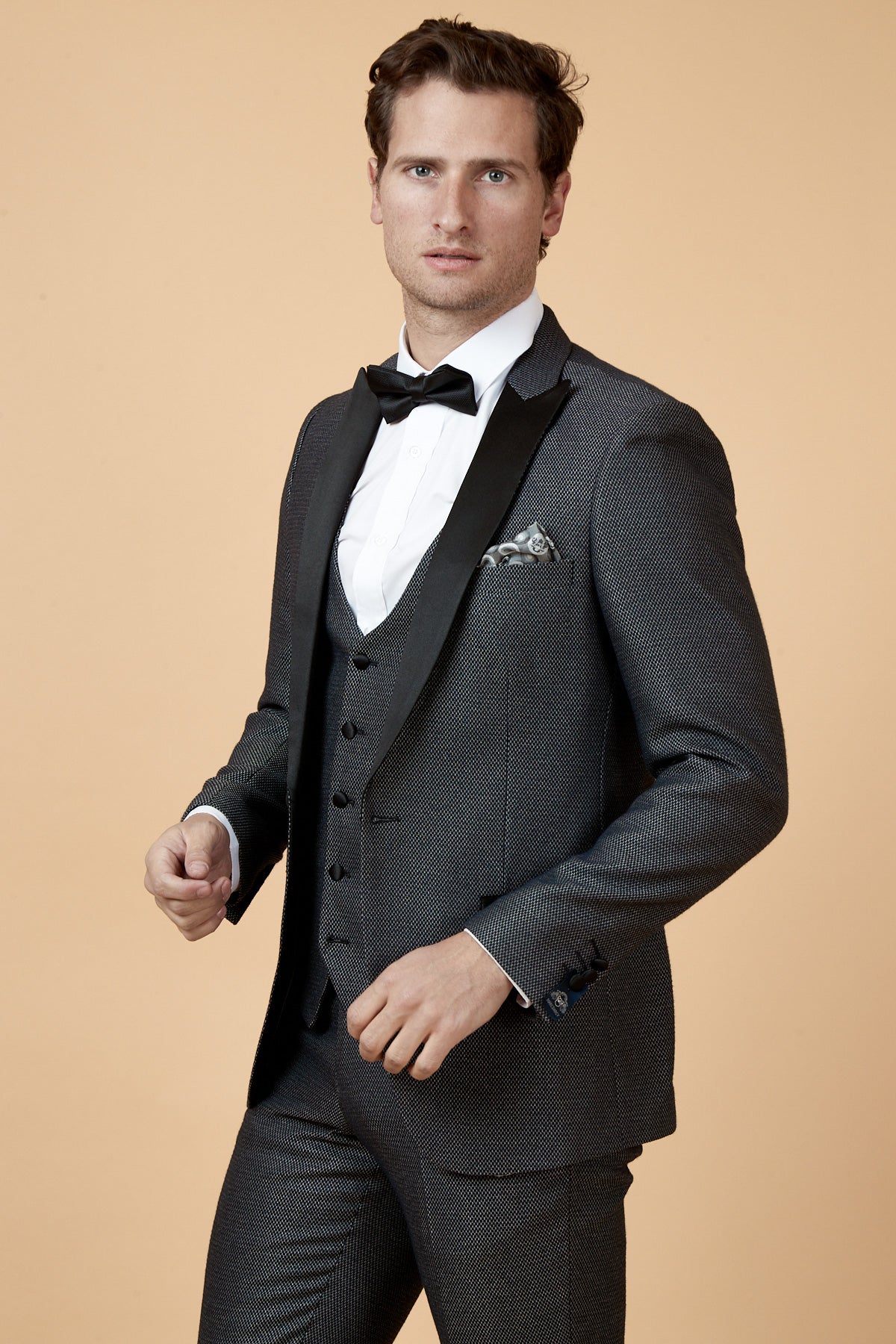 A man wears the marcdarcy SPENCER Charcoal Grey Tux Lapel Blazer with a black satin lapel, white shirt, black bow tie, and pocket square against a beige background—ideal for black tie events.